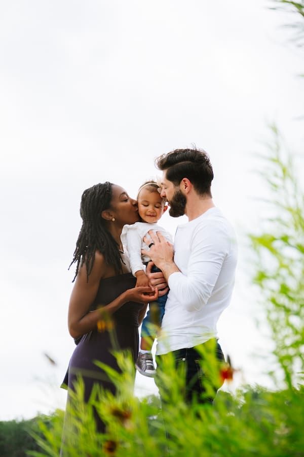 Interracial family with mother, father, and toddler sharing a tender moment outdoors among green plants.