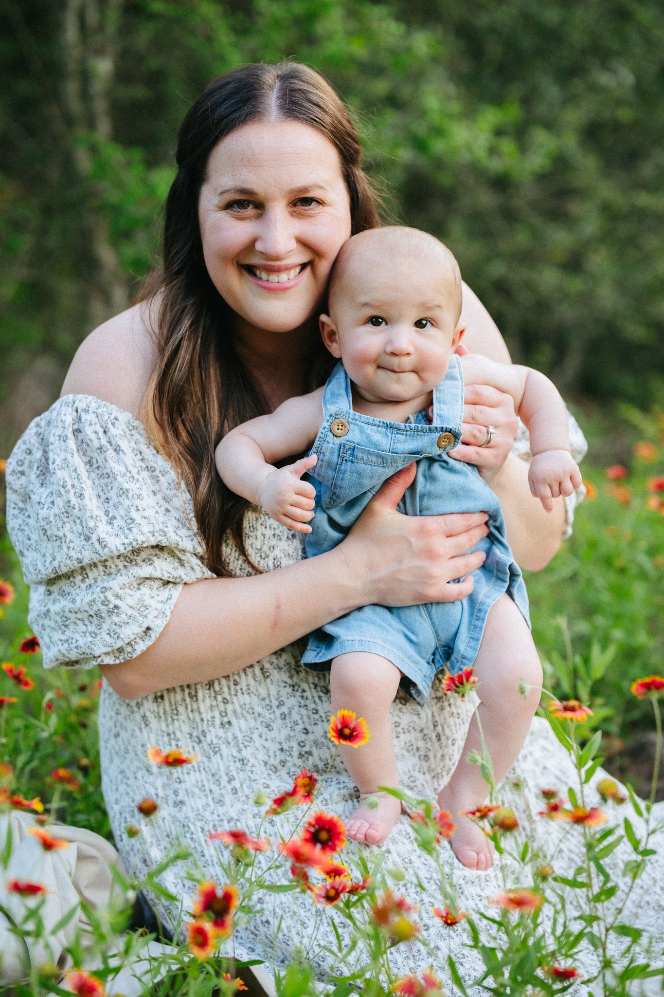 Smiling mother holding baby in denim overalls among red wildflowers in a lush green outdoor setting.