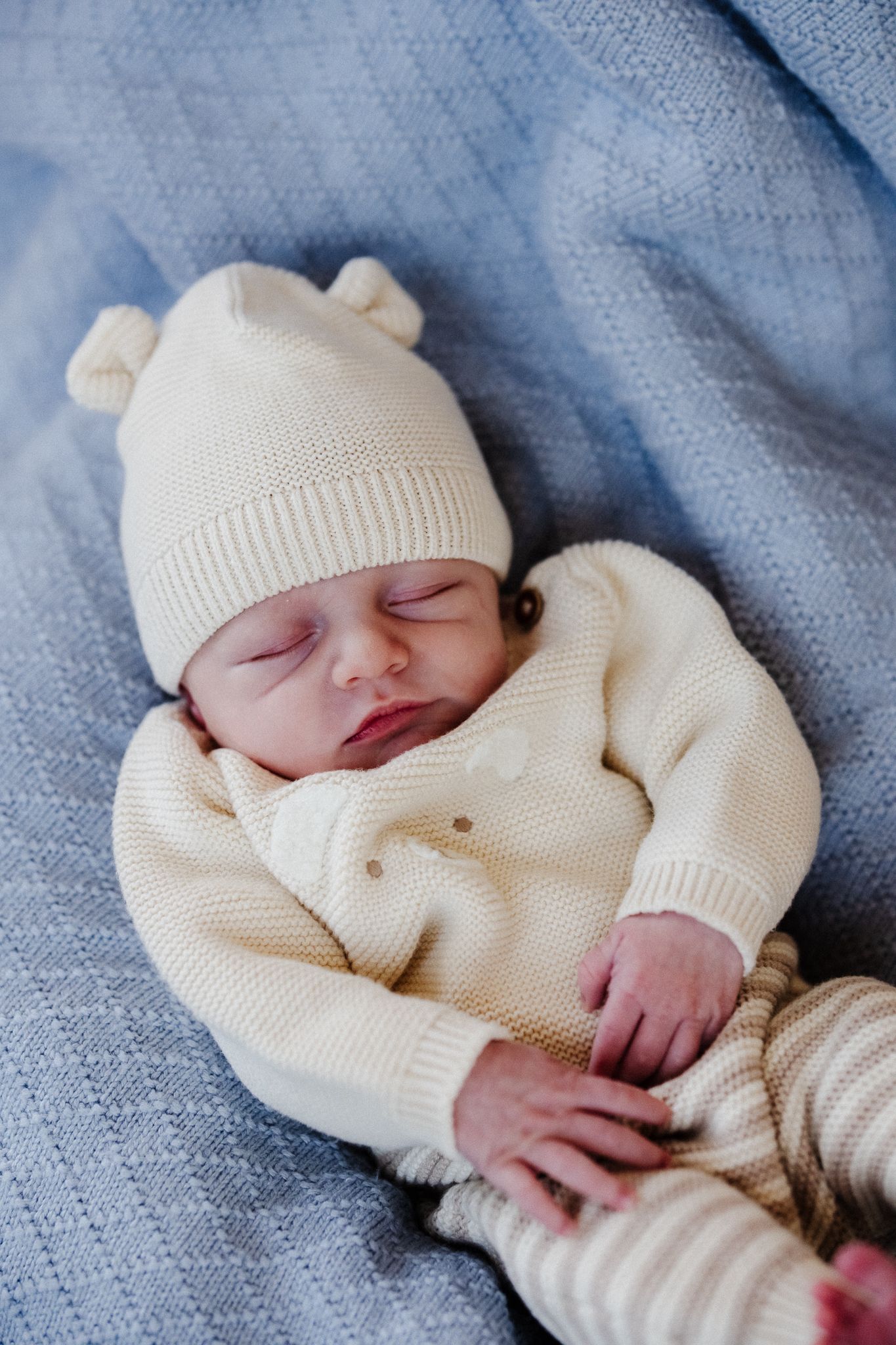A sleeping newborn baby wearing a cozy cream-colored knit sweater and matching hat lies on a textured blue blanket.
