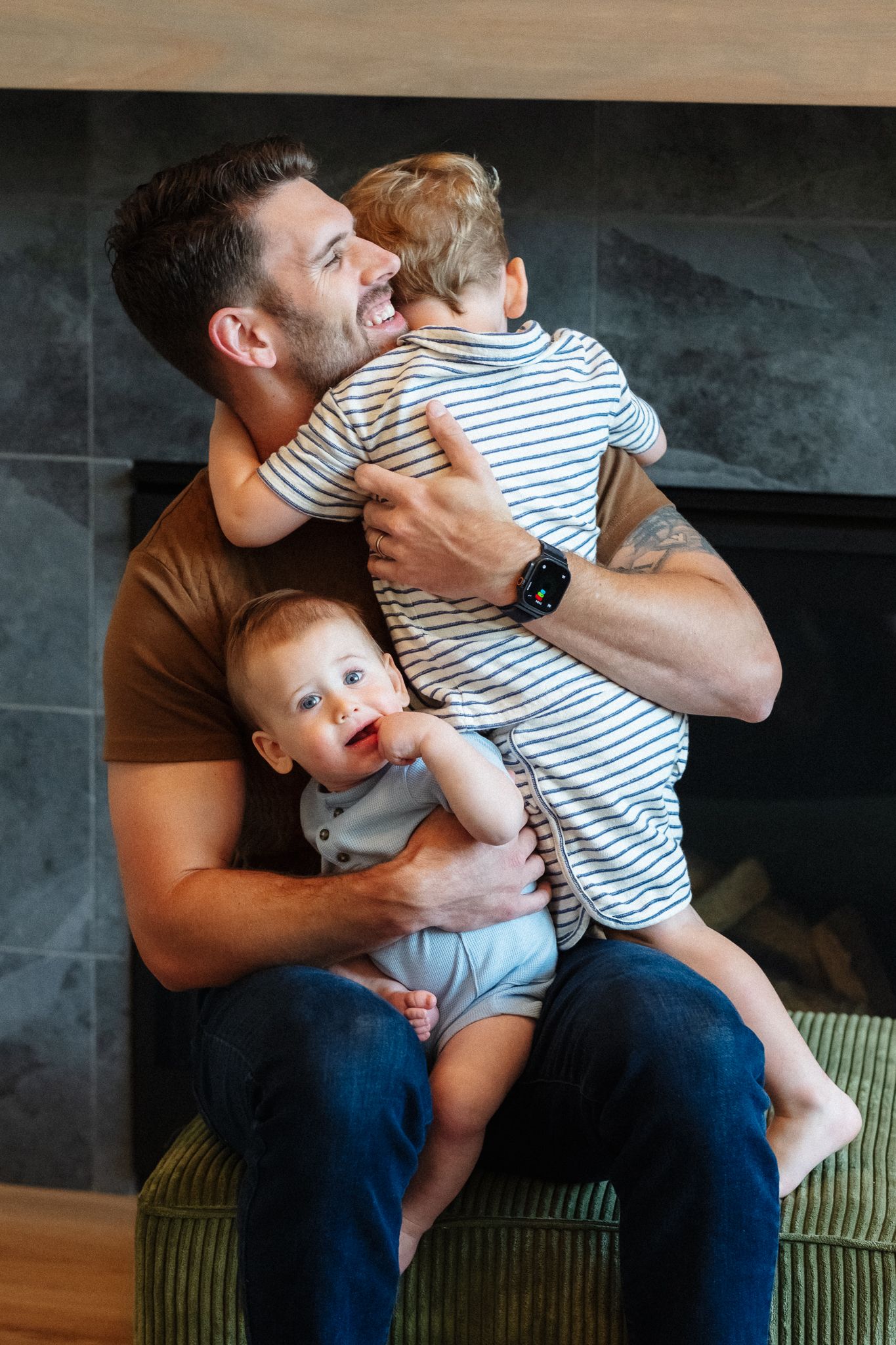 A smiling father holds two young children, a toddler and a baby, in front of a modern fireplace at home.