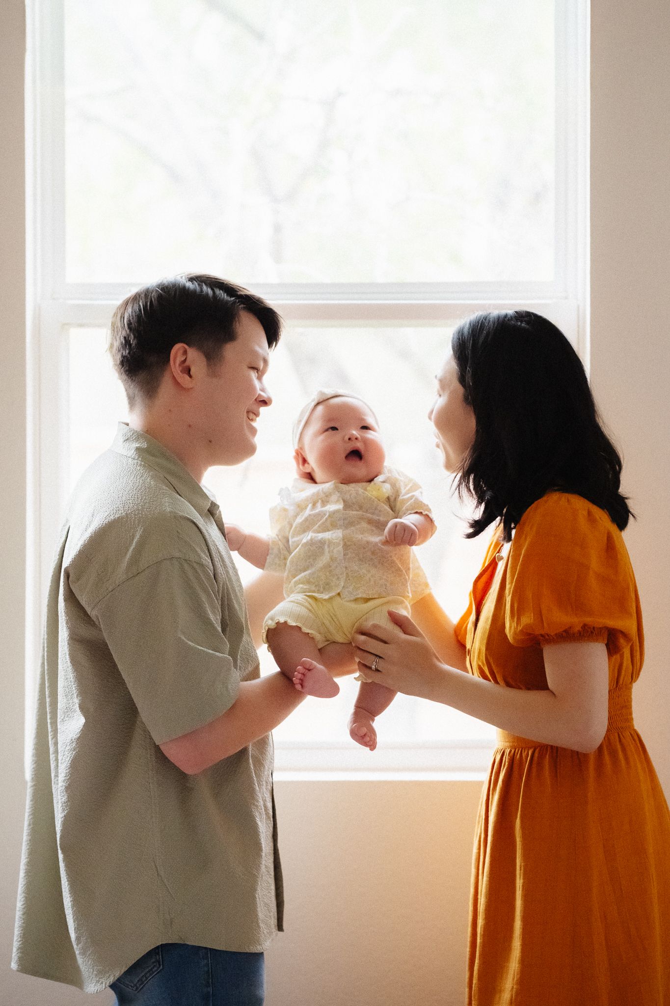 Happy parents holding their smiling baby in a bright, sunlit room, sharing a joyful family moment.