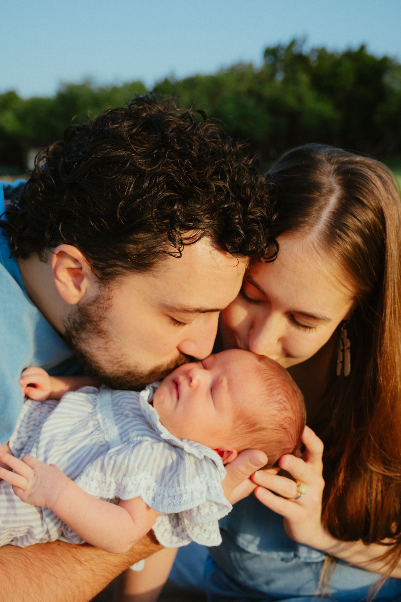 Parents gently kiss their sleeping newborn baby outdoors during golden hour light.