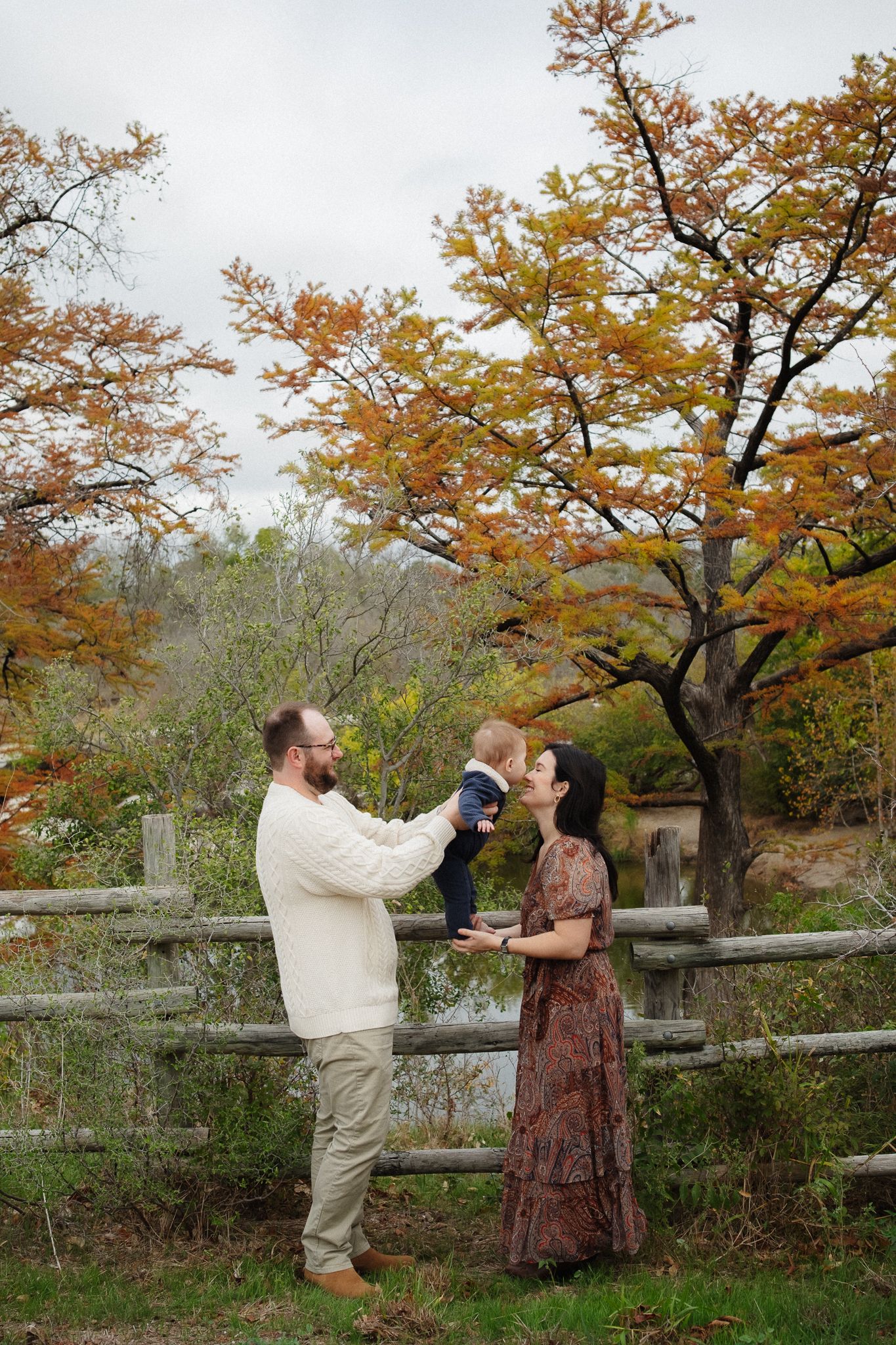 A couple shares a romantic moment under autumn trees with orange foliage near a wooden fence.