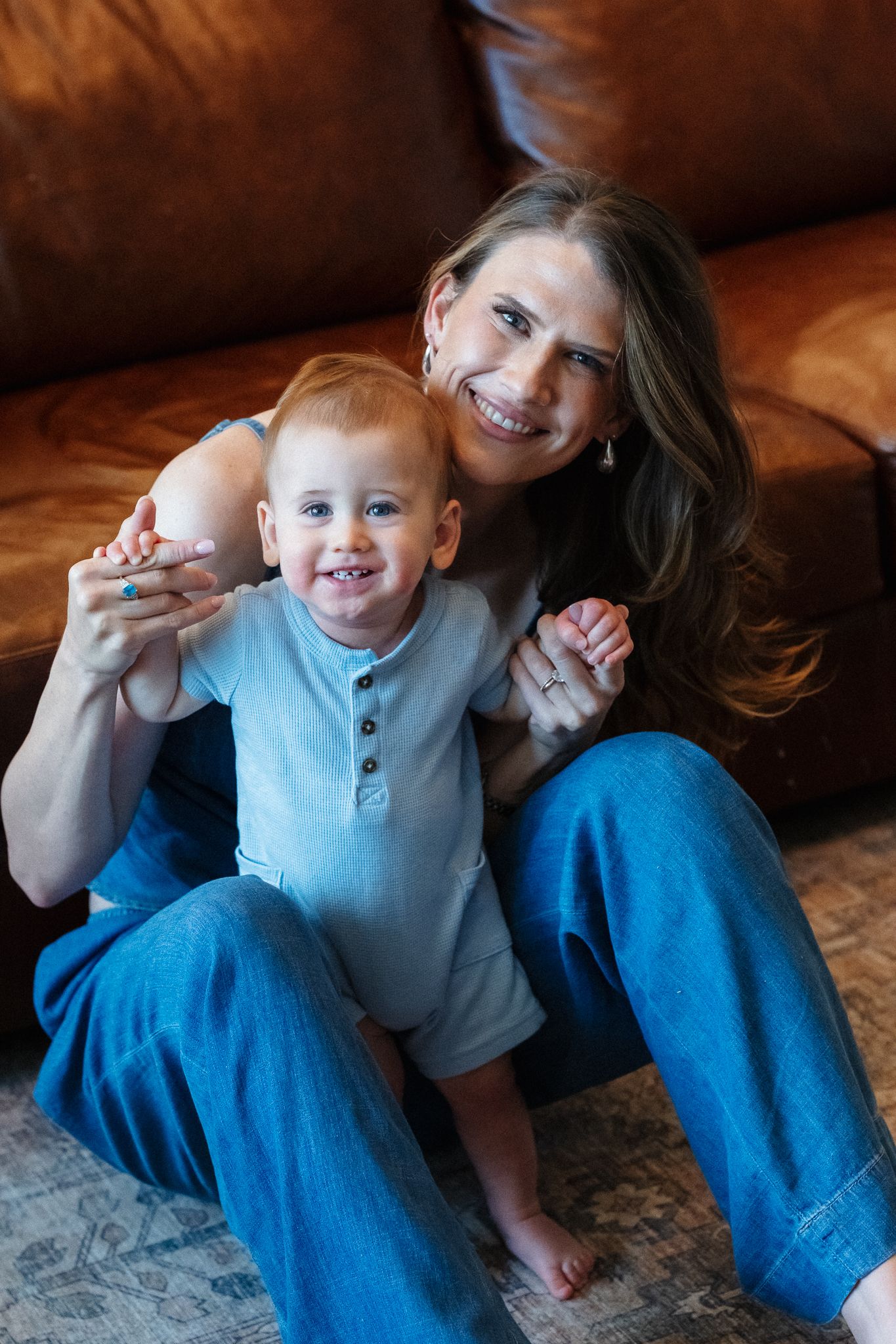 A smiling woman sits on the floor against a leather sofa, hugging a happy red-haired toddler boy in a blue outfit.