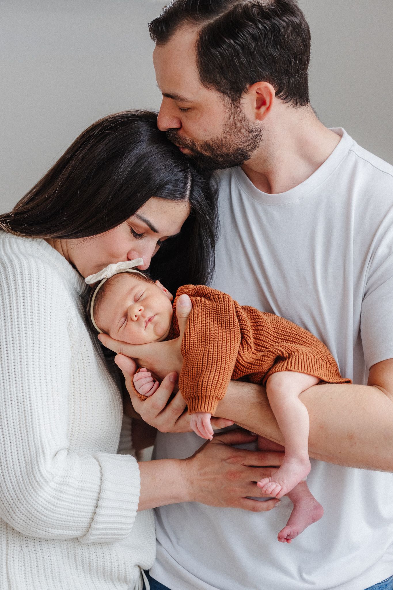 New parents kissing their sleeping newborn baby dressed in orange knit outfit against gray background.