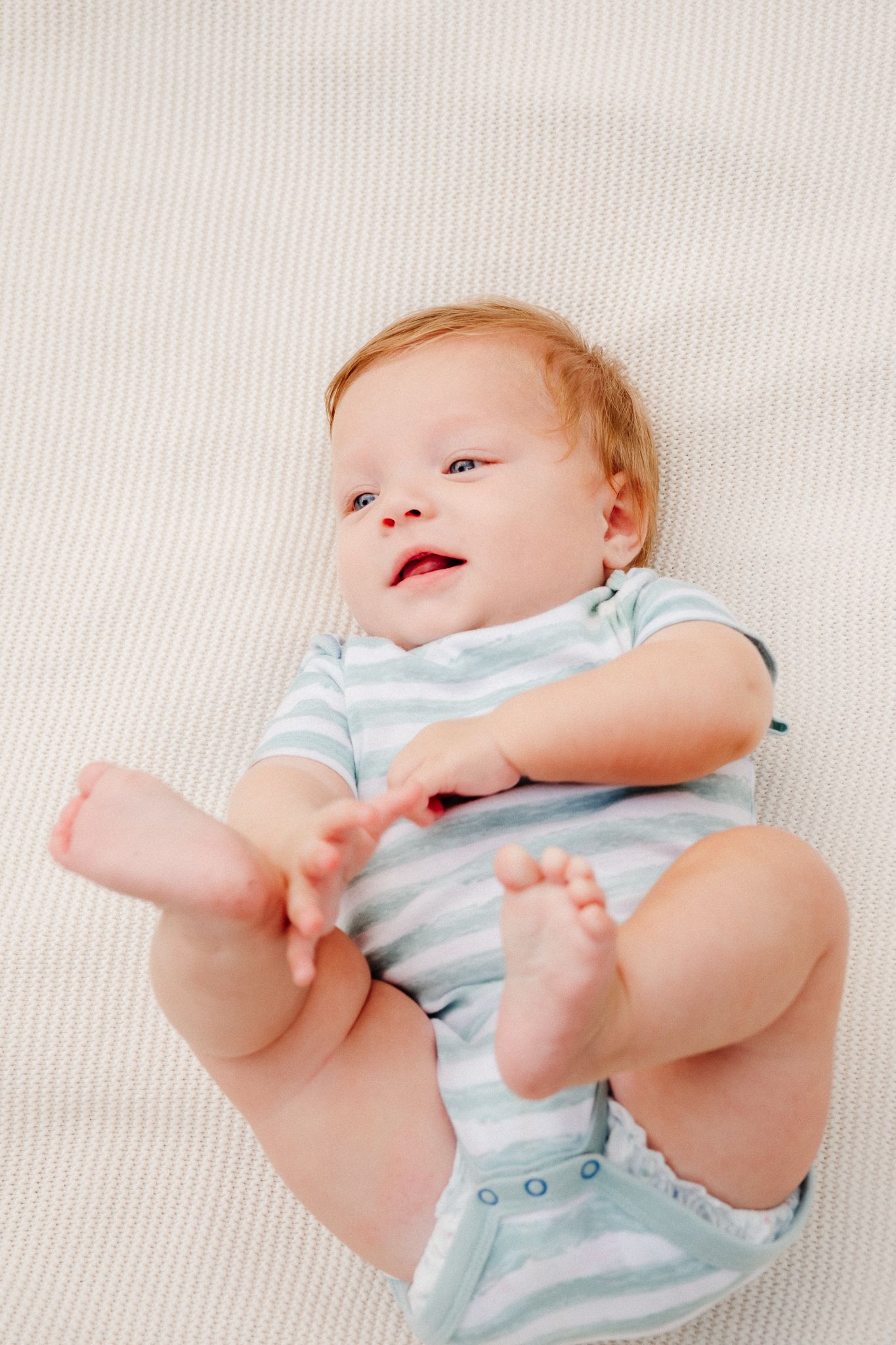 Baby wearing light blue and white striped onesie lying on white blanket with feet in focus.