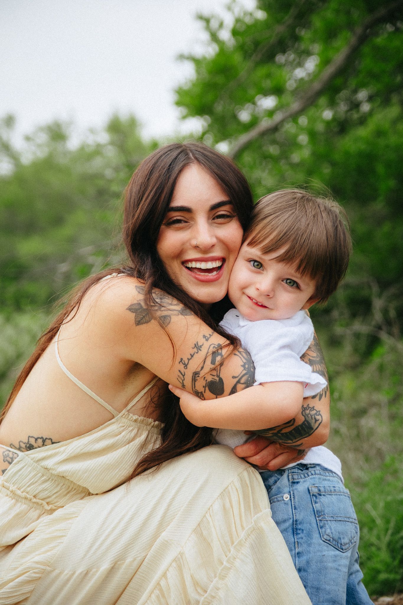 A parent and young child share a joyful embrace outdoors among green foliage on a sunny day.