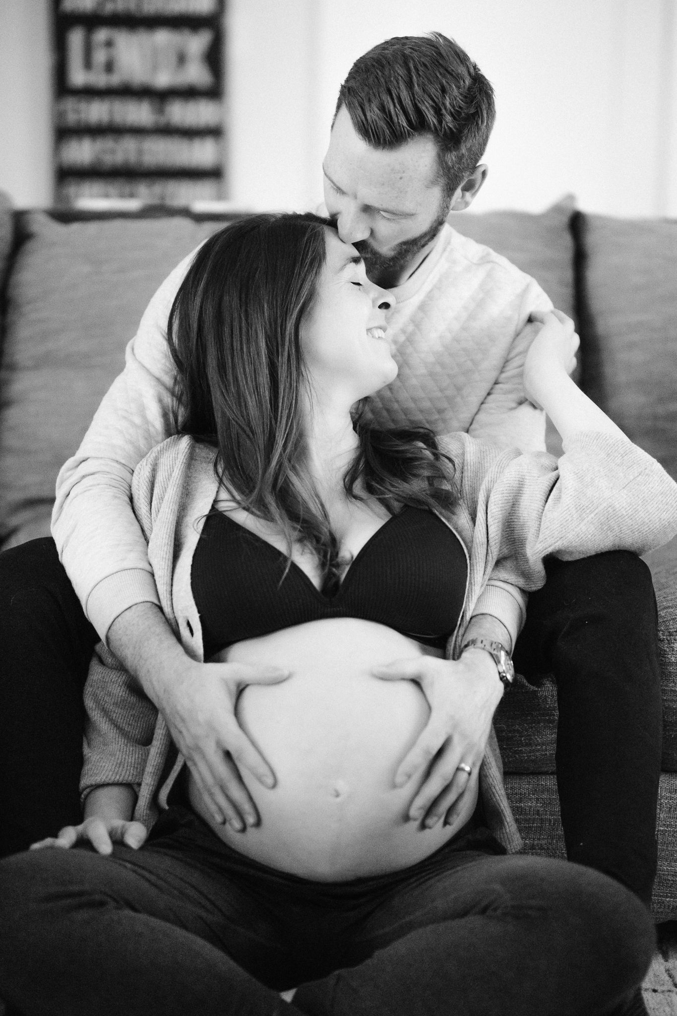 A couple shares an intimate moment on a couch while cradling a pregnant belly in black and white.