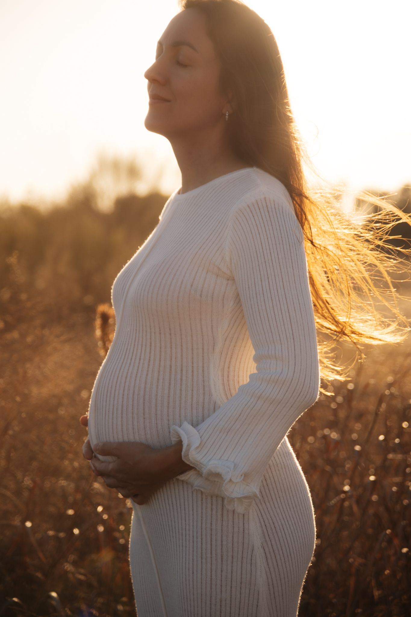Pregnant woman in white sweater dress standing in golden sunset light with flowing hair in nature.