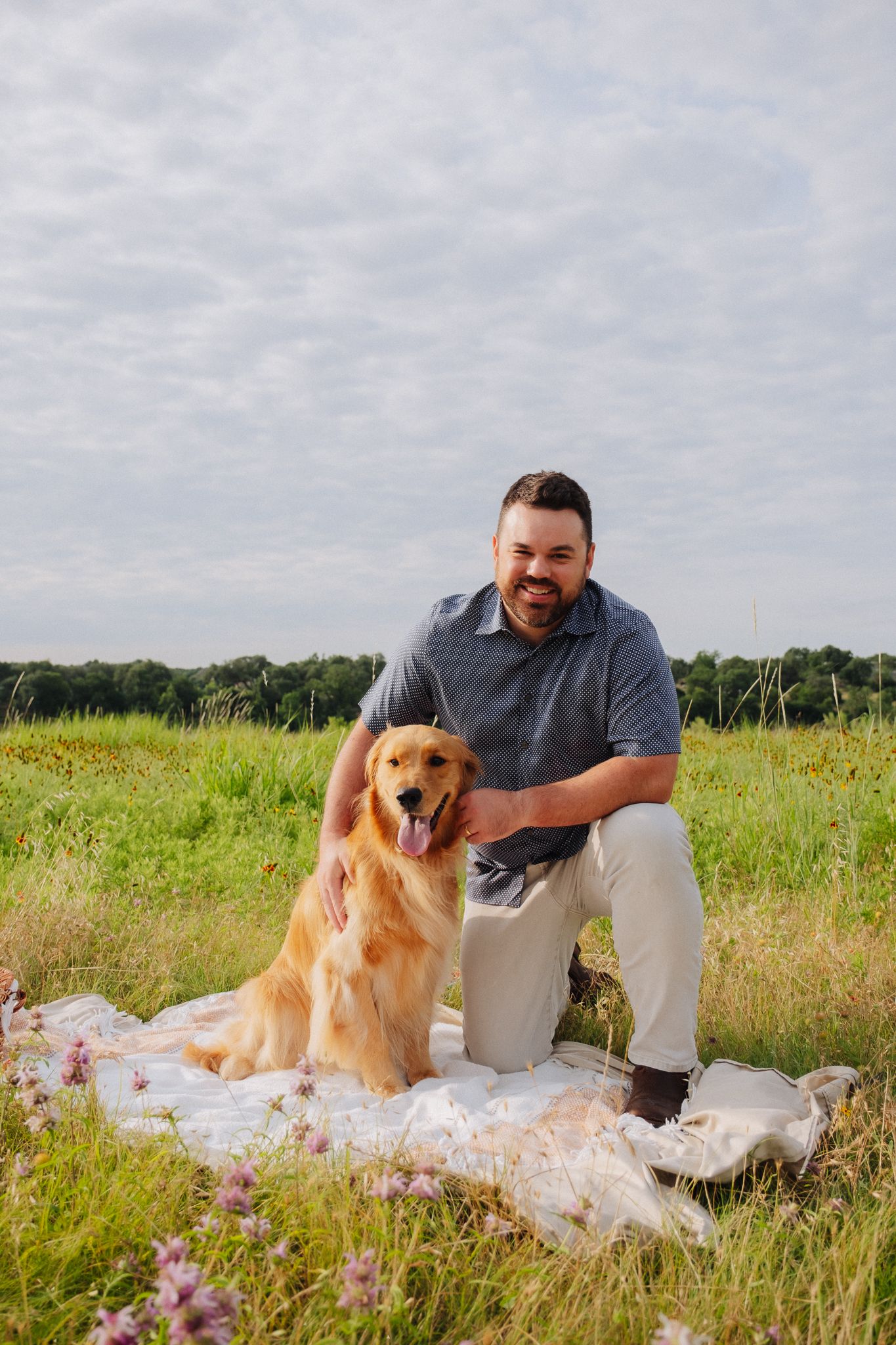 Golden retriever poses with their companion on a white blanket in a wildflower meadow on a cloudy day.