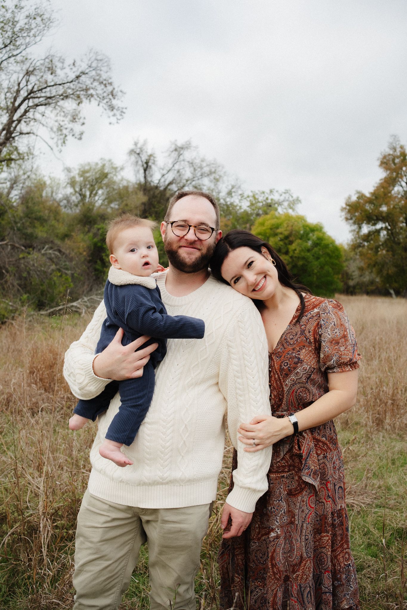A heartwarming family portrait taken in a natural field setting during autumn with trees in the background.