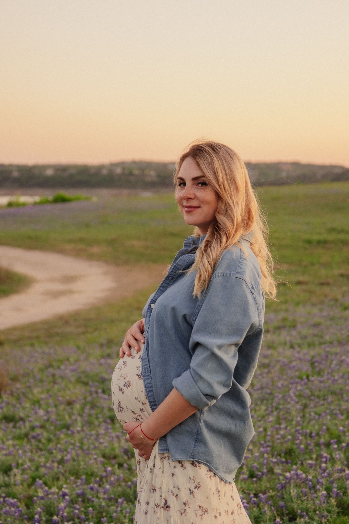 A pregnant woman in a denim jacket stands in a field of purple wildflowers at sunset.