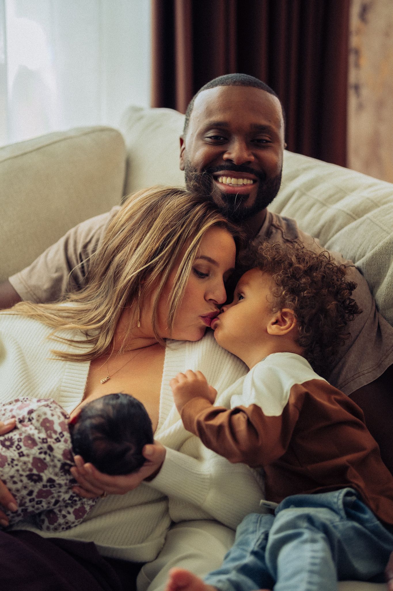 Happy interracial family sitting on couch, parents kissing toddler while holding newborn baby at home.