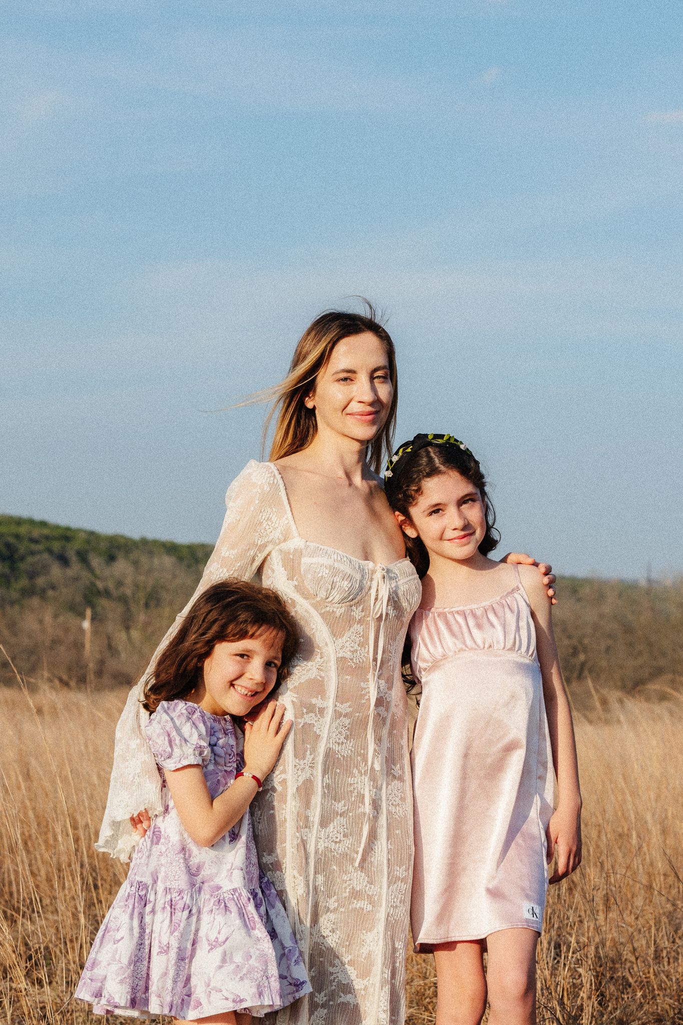 A woman in a white lace dress poses with two young girls in a sunny field under a blue sky.