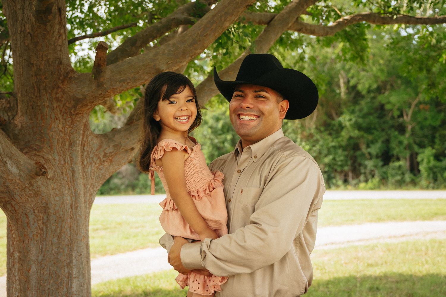 A smiling man in a black cowboy hat holds a young girl in a pink dress outdoors near a large tree.