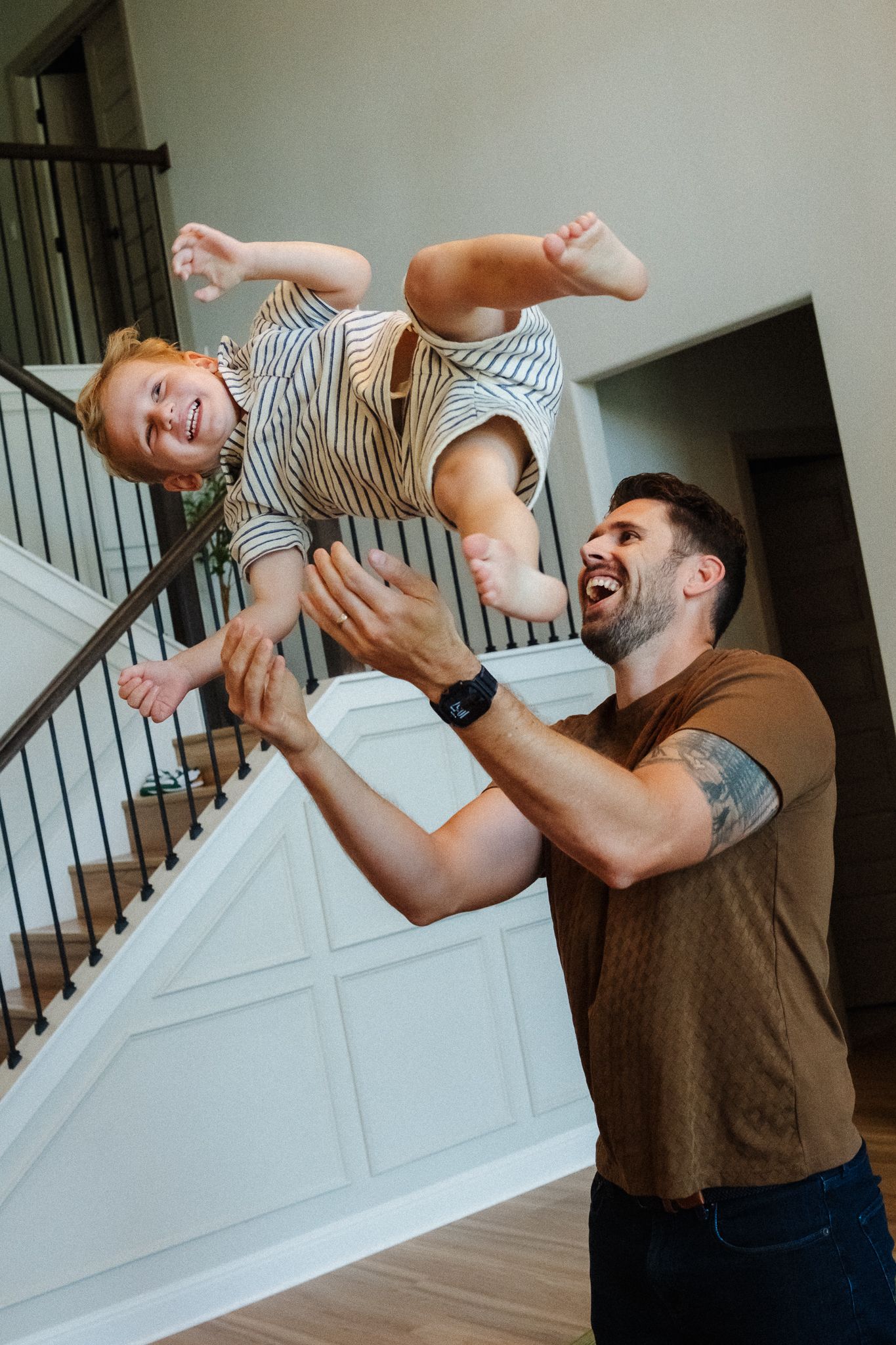 A laughing father lifts his giggling redhead toddler high in the air inside a modern home with a staircase.