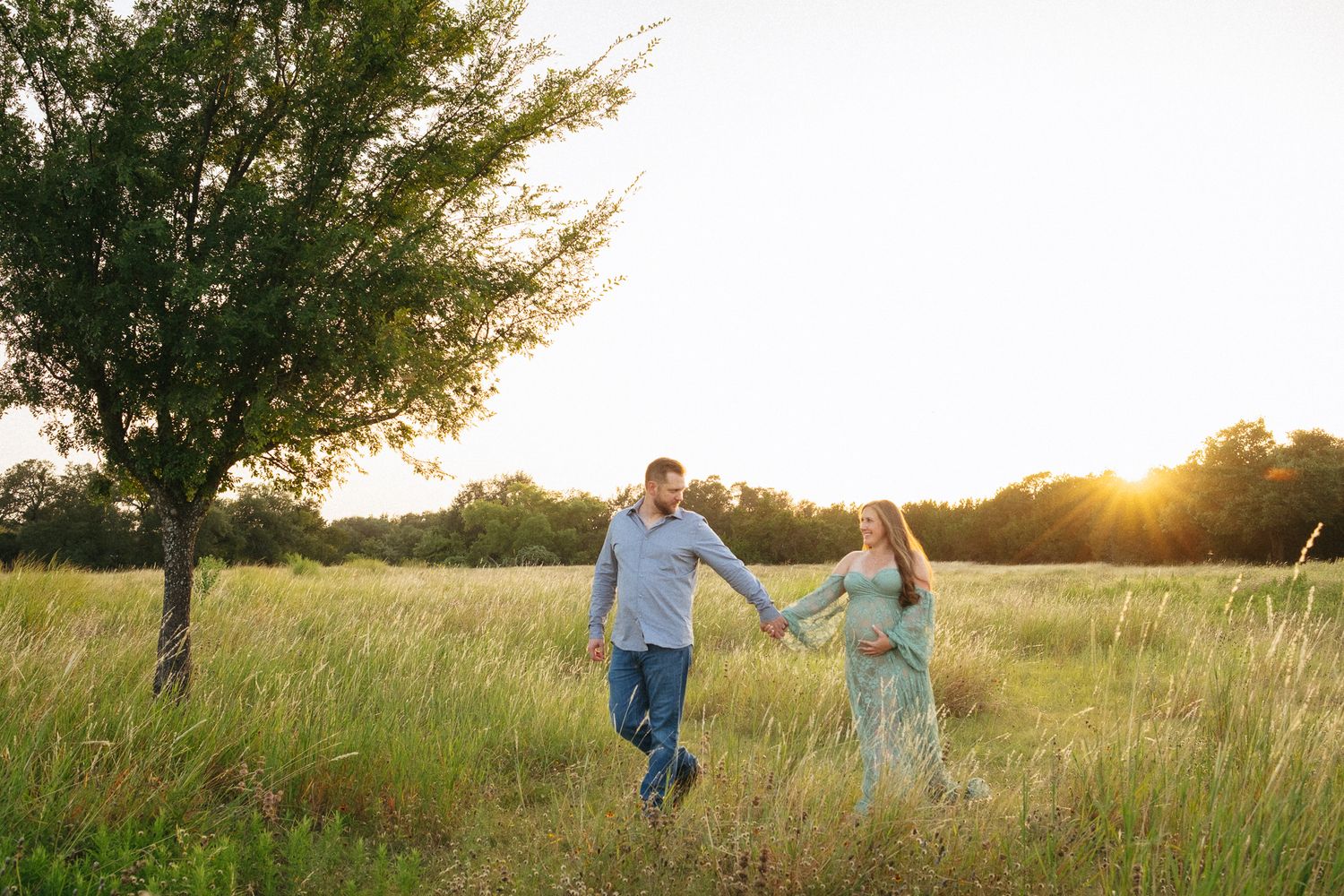 Couple holding hands walking through golden sunlit meadow with tall grass and trees at sunset.