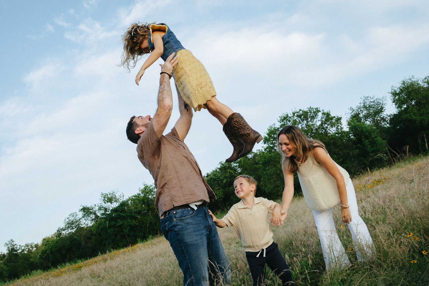 Family playing outdoors in a meadow, father lifting child in the air while mother and son watch joyfully.