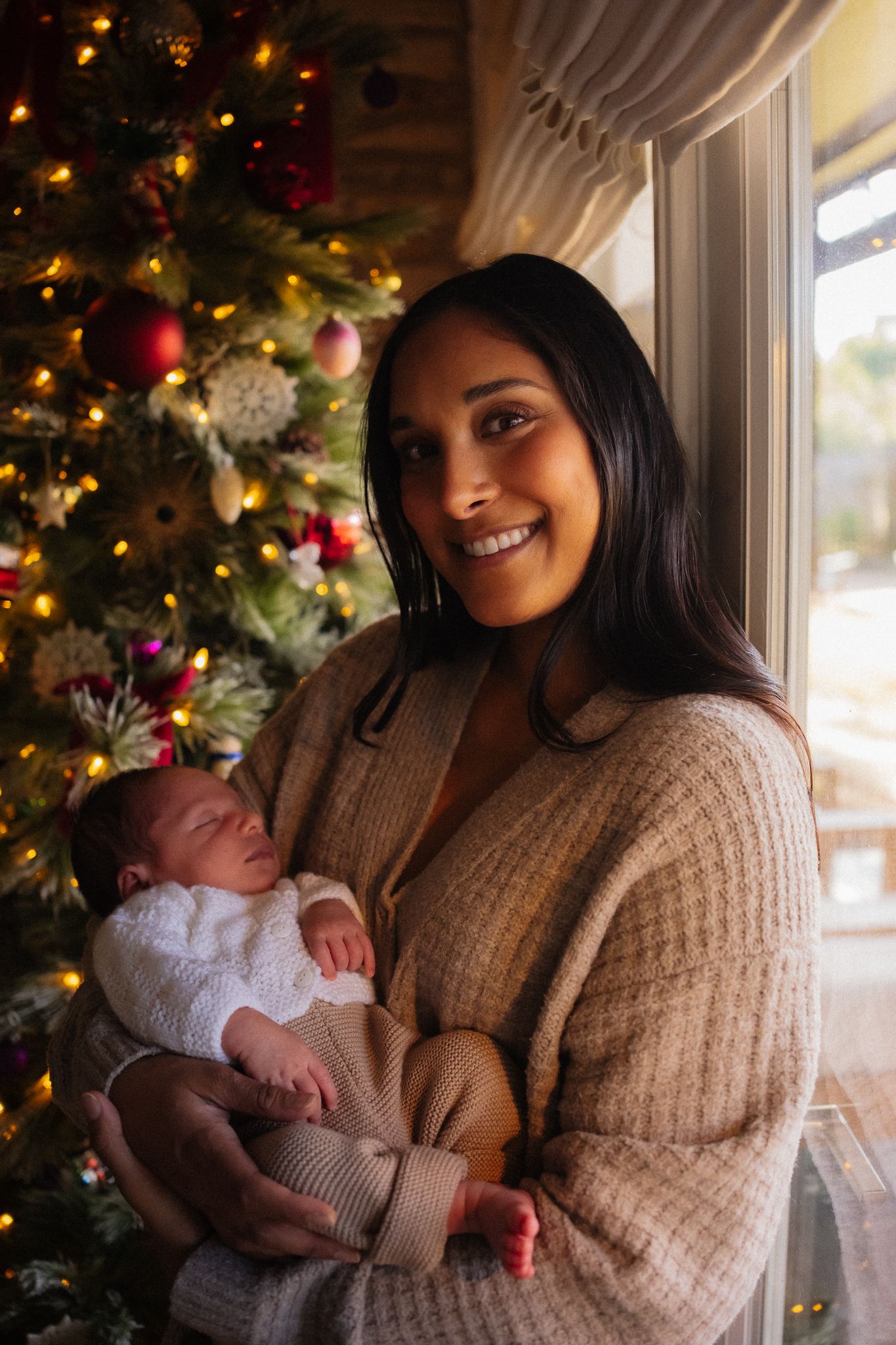 Smiling mother holding newborn baby in front of decorated Christmas tree with warm glowing lights.