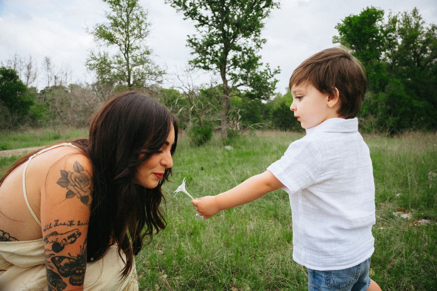Tattooed mother bends down as young boy offers her a dandelion in a lush green field.