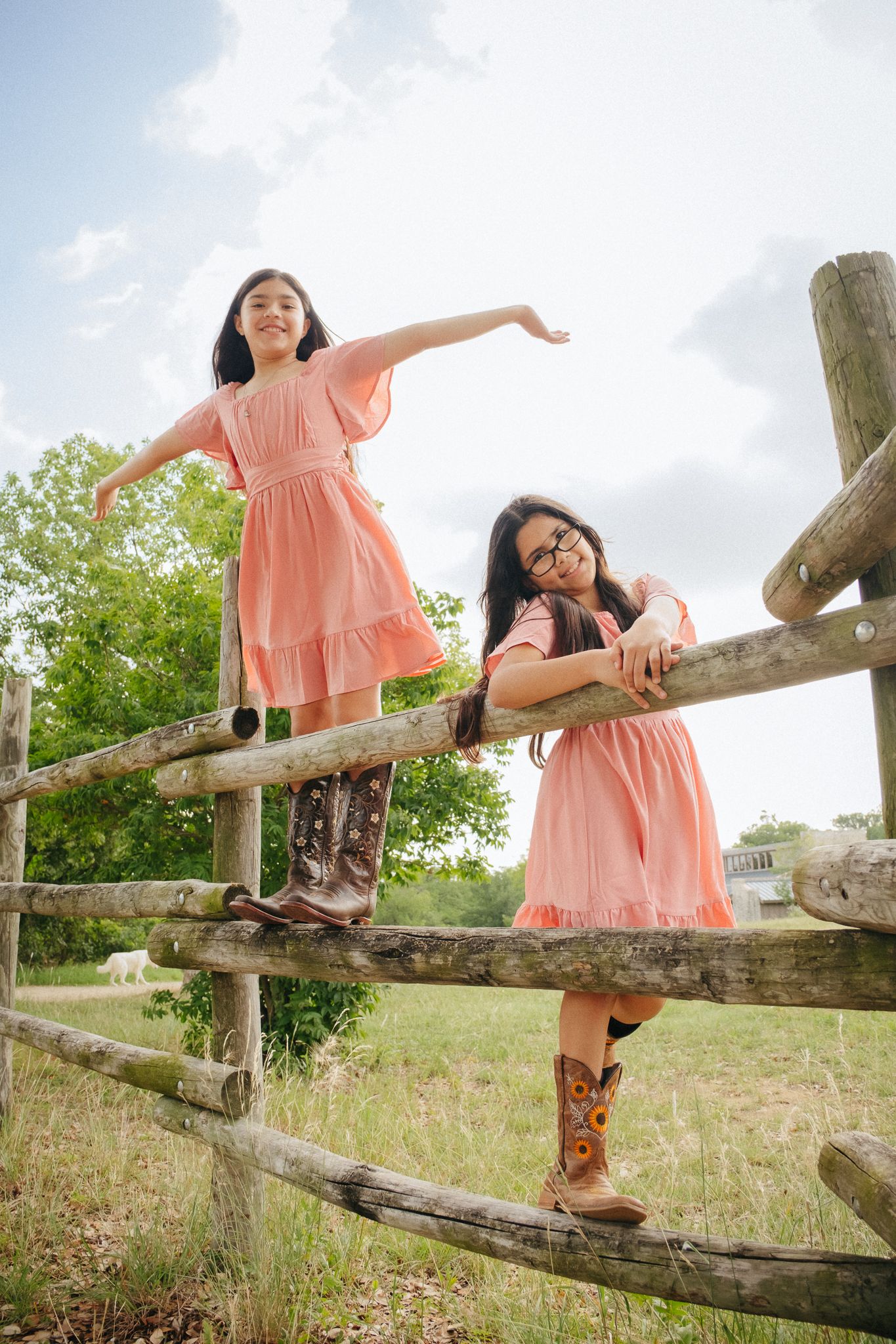 Young people in salmon-colored dresses playfully pose along a rustic wooden fence in a sunny field.