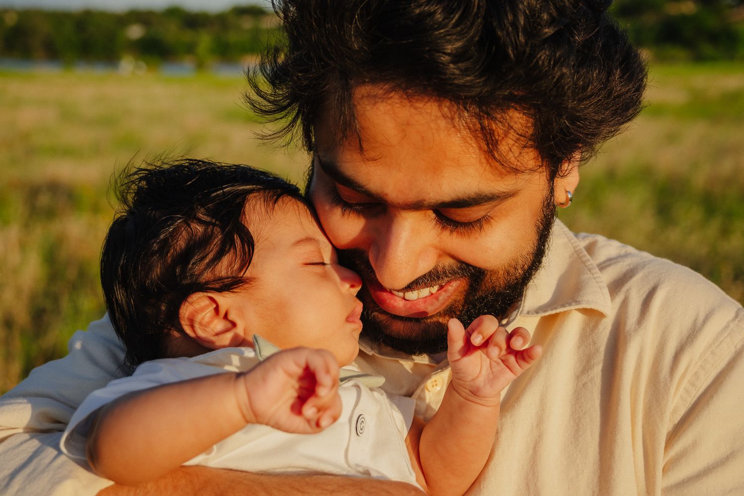 A tender moment between a parent and baby is captured in warm golden sunlight in an outdoor field setting.