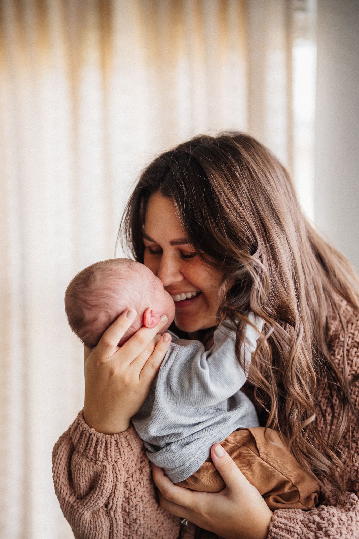 Smiling mother with long brown hair tenderly kissing her newborn baby wrapped in a gray onesie.