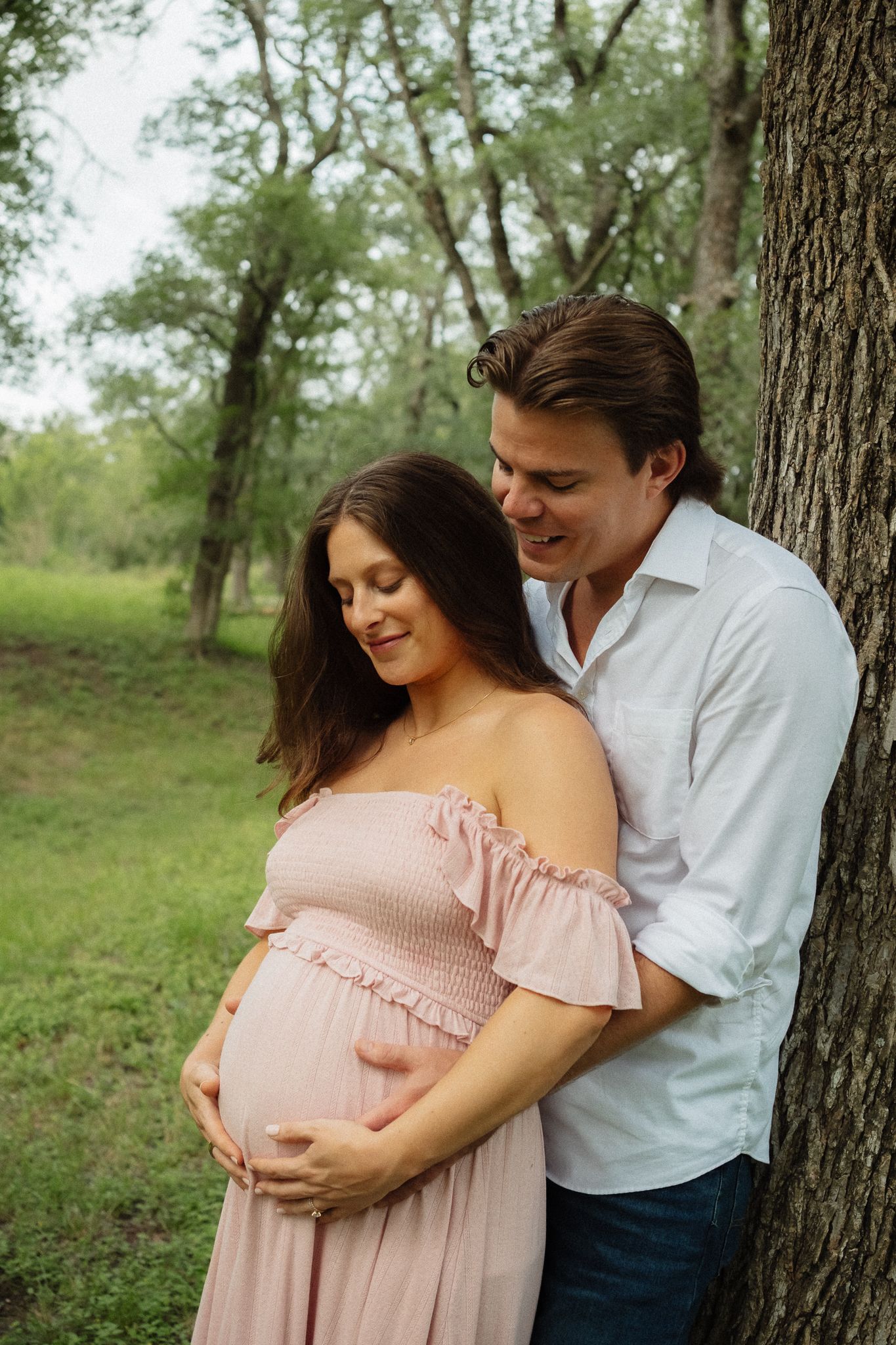 Couple sharing a tender maternity moment outdoors, woman in pink off-shoulder dress cradling baby bump against a tree.