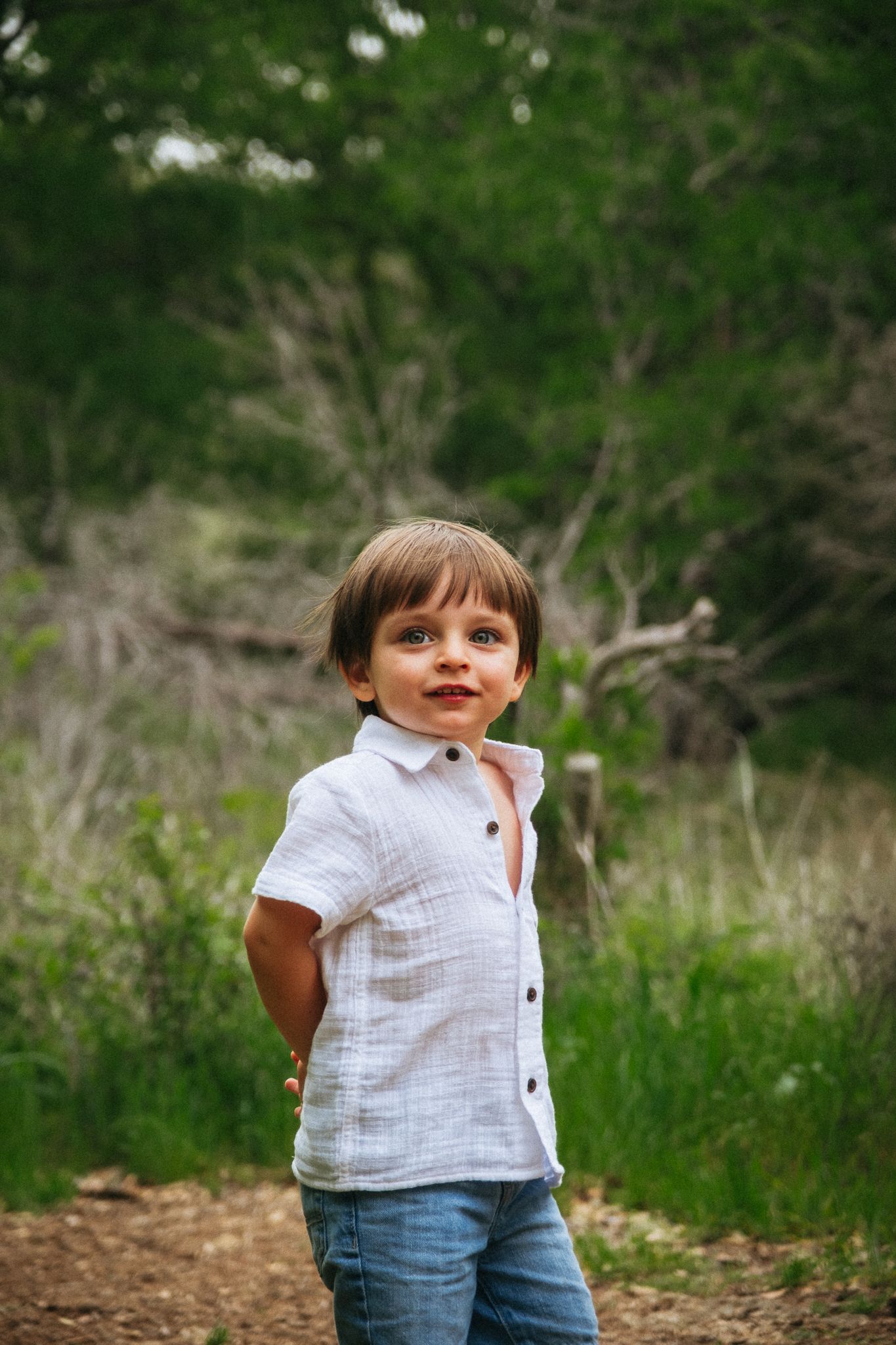 A toddler in a white shirt and jeans stands playfully in a natural outdoor setting with greenery in the background.