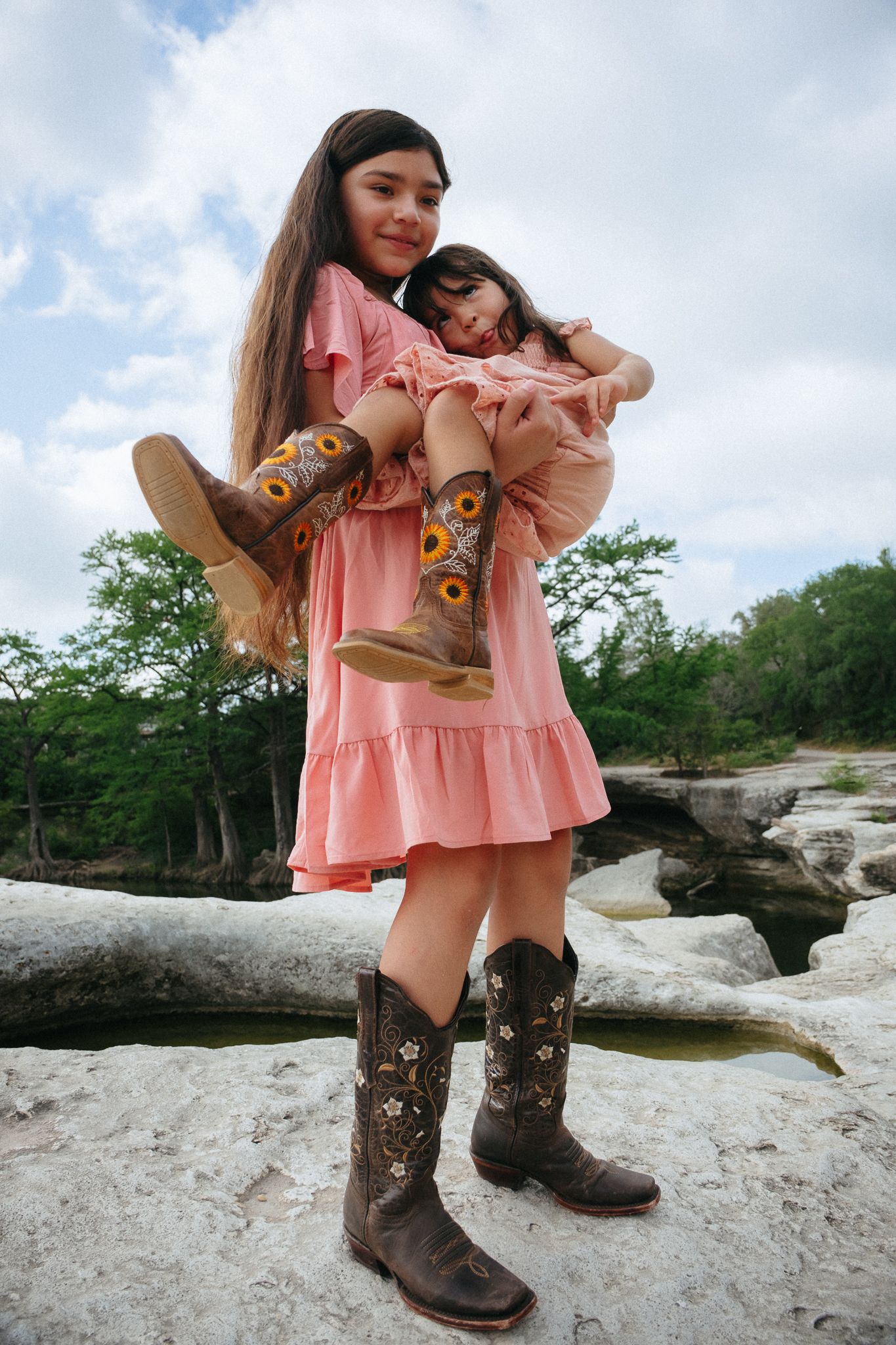 Two people in matching pink dresses and cowboy boots pose together on rocky terrain against a cloudy sky background.