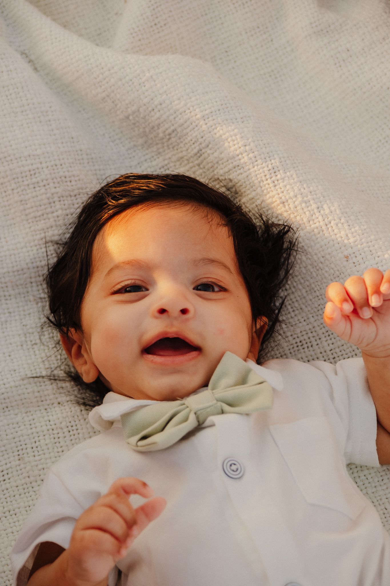 A small baby wearing a white outfit with a bow collar lies on a soft gray blanket smiling joyfully.