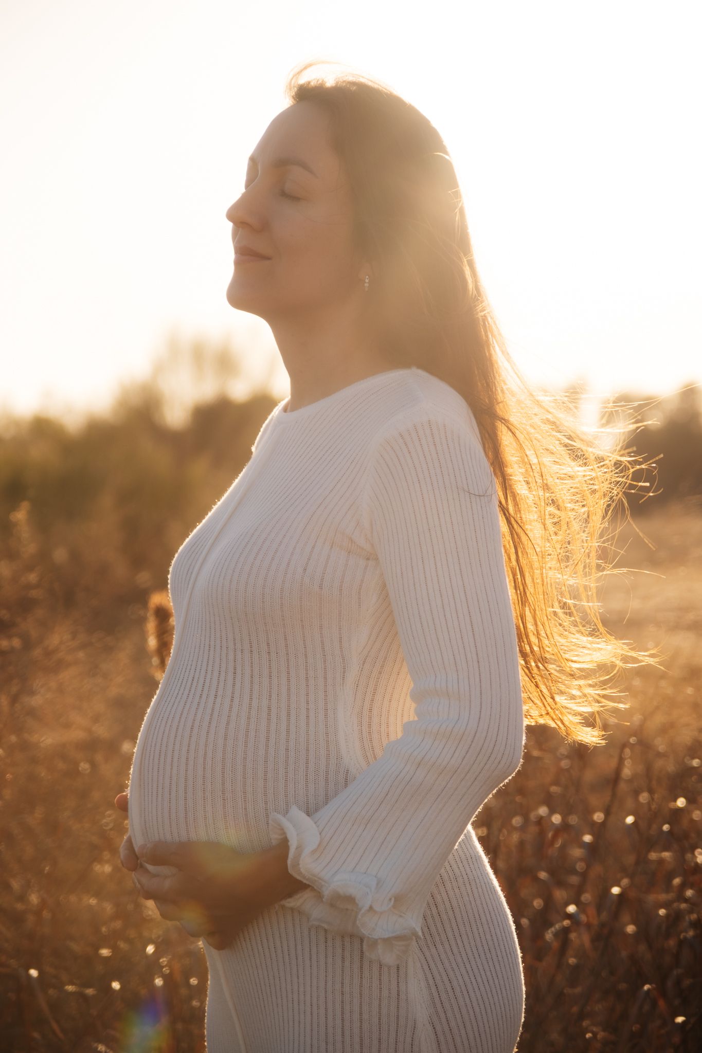A pregnant woman in a white sweater dress stands in a sunlit field at golden hour with her hair flowing in the breeze.
