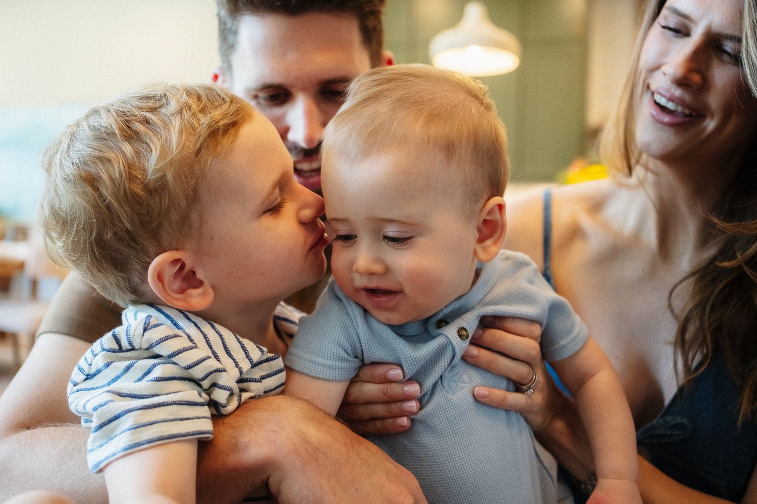 Happy family with parents and two young boys, older sibling kissing baby brother on the cheek indoors.