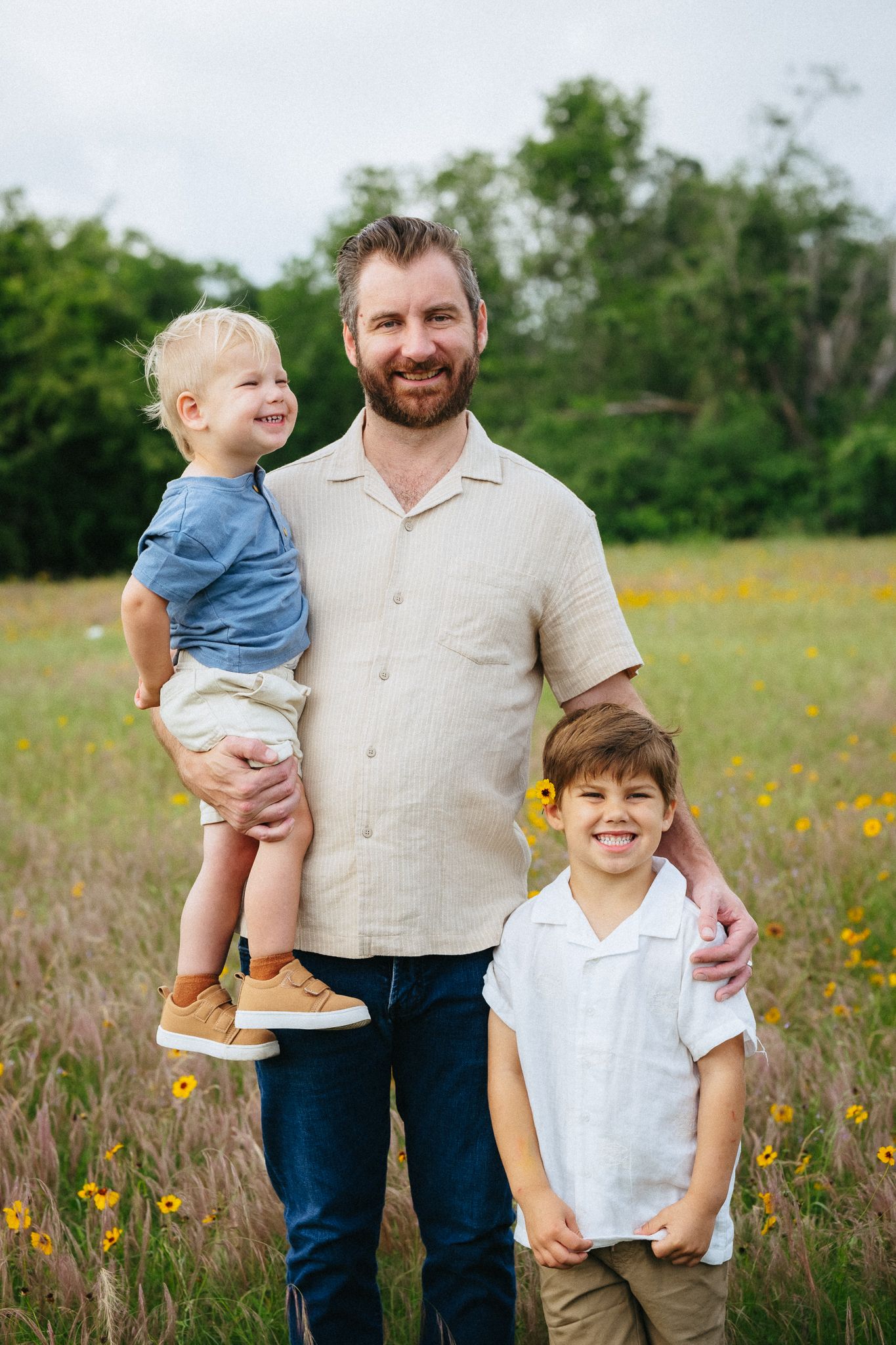 A bearded father stands in a wildflower field holding a toddler while his older son smiles beside him.