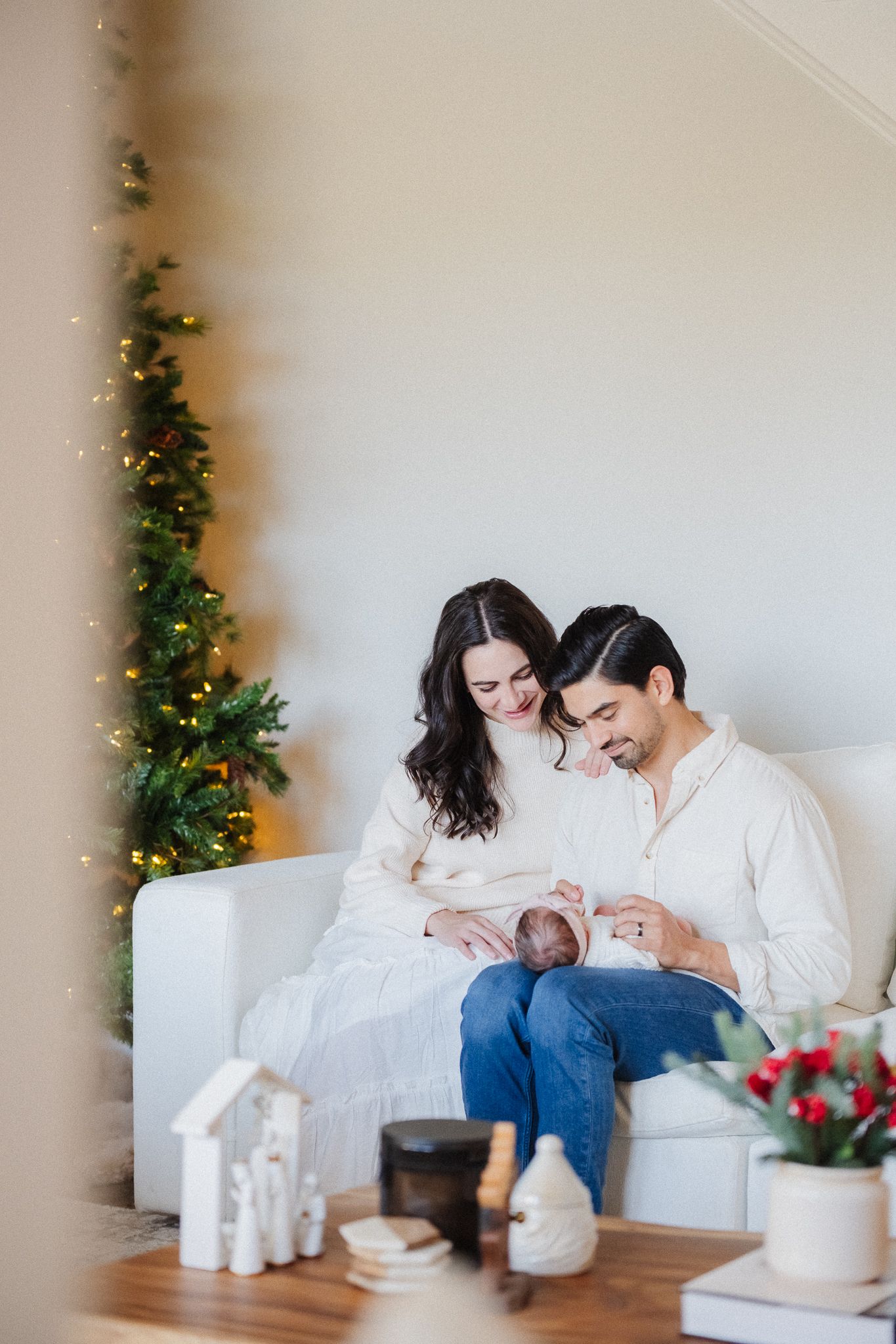 A couple in white sweaters sits on a sofa holding their newborn baby beside a decorated Christmas tree.