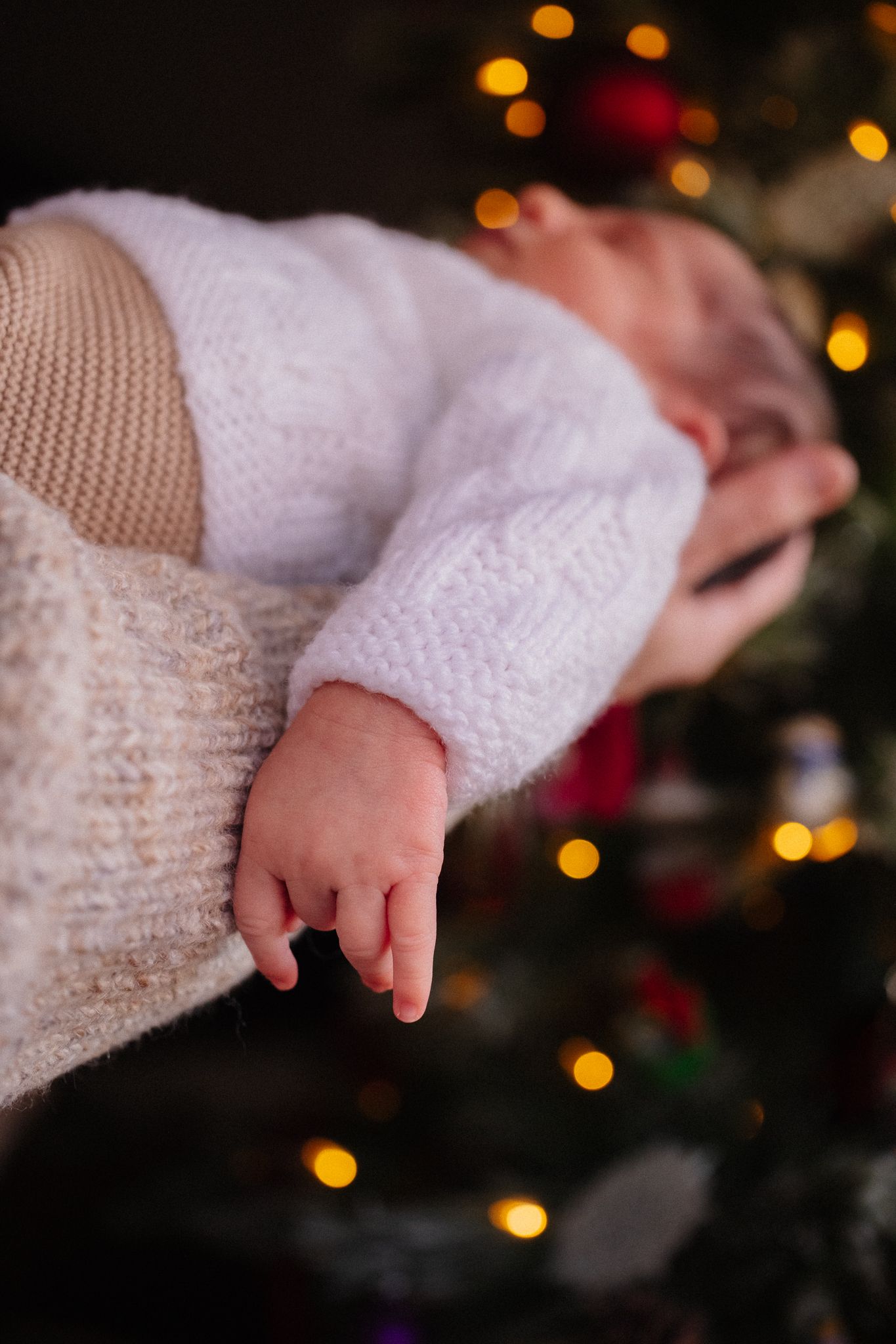 Newborn baby in white knit outfit cradled in arms near a glowing Christmas tree with bokeh lights.