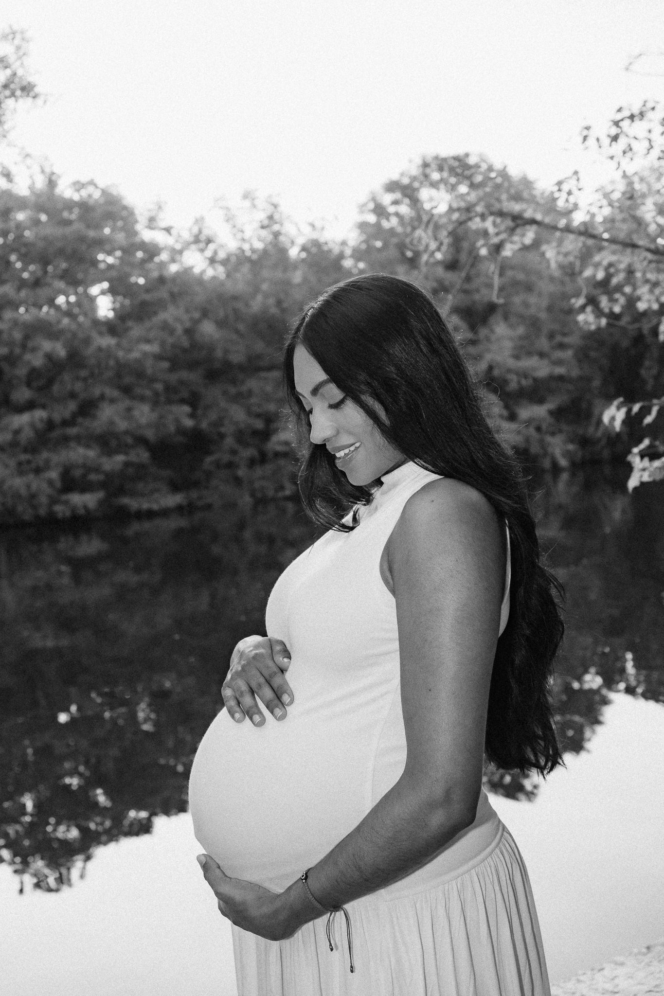 Black and white maternity photo of a smiling pregnant woman cradling her belly outdoors near a reflective pond.
