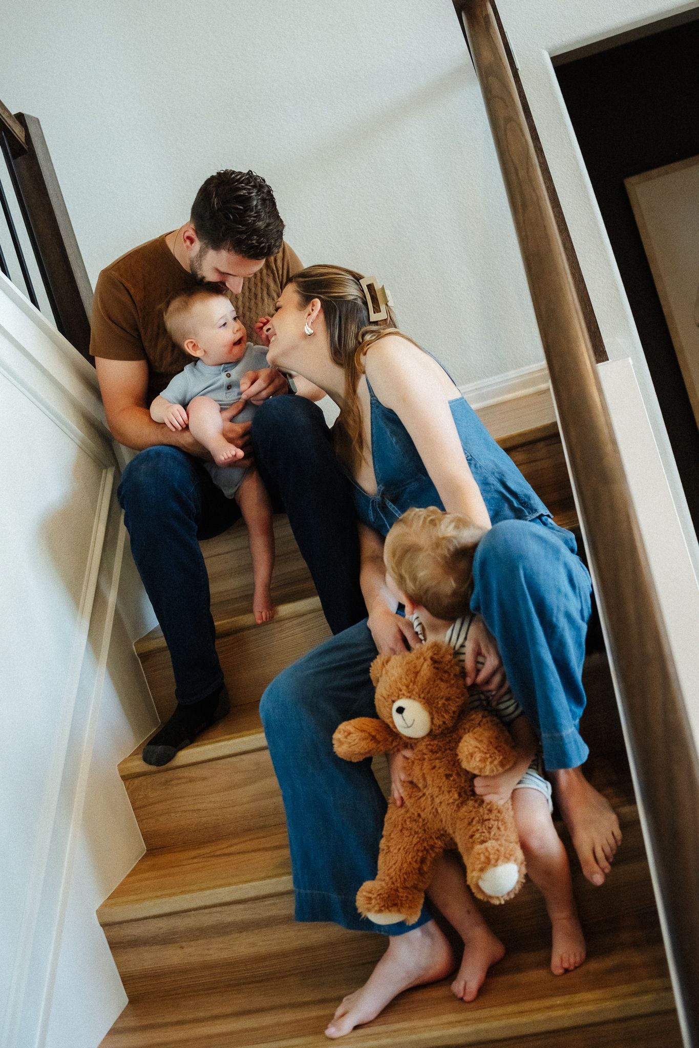 Family of four sitting on wooden stairs, parents kissing baby while toddler hugs teddy bear.