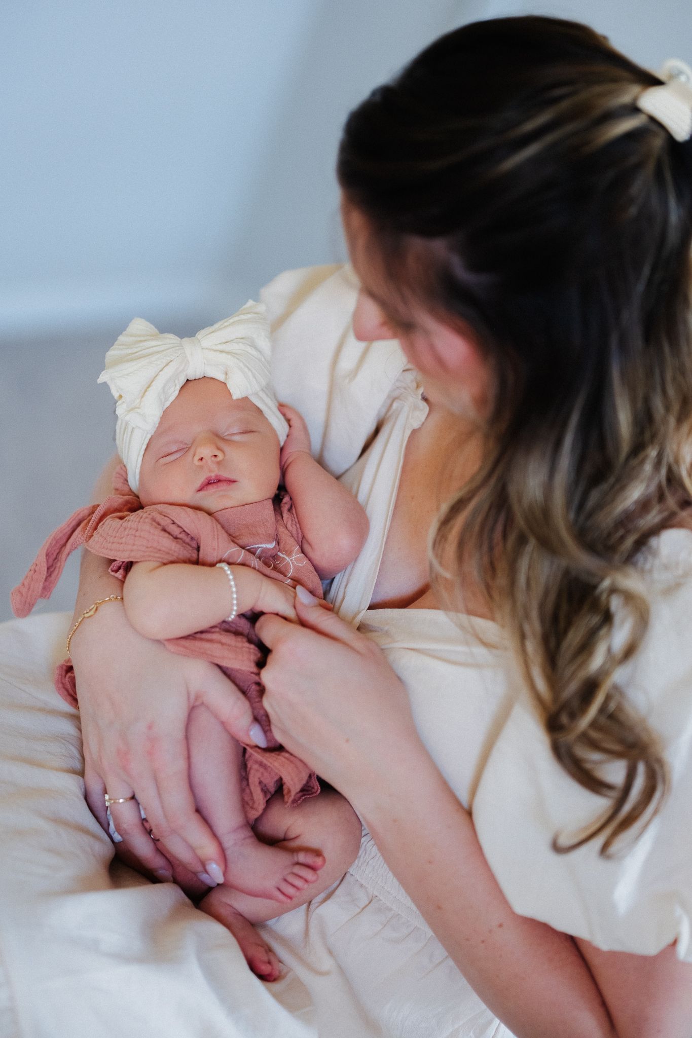 Mother in white dress gazes lovingly at sleeping newborn baby wrapped in pink blanket with white bow headband.