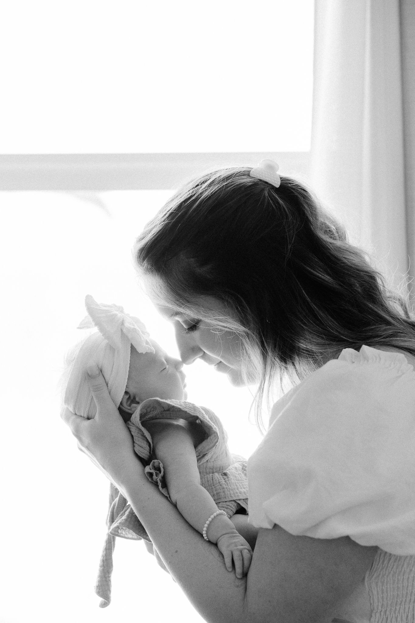 Black and white photo of a mother tenderly holding her newborn baby near a bright window.