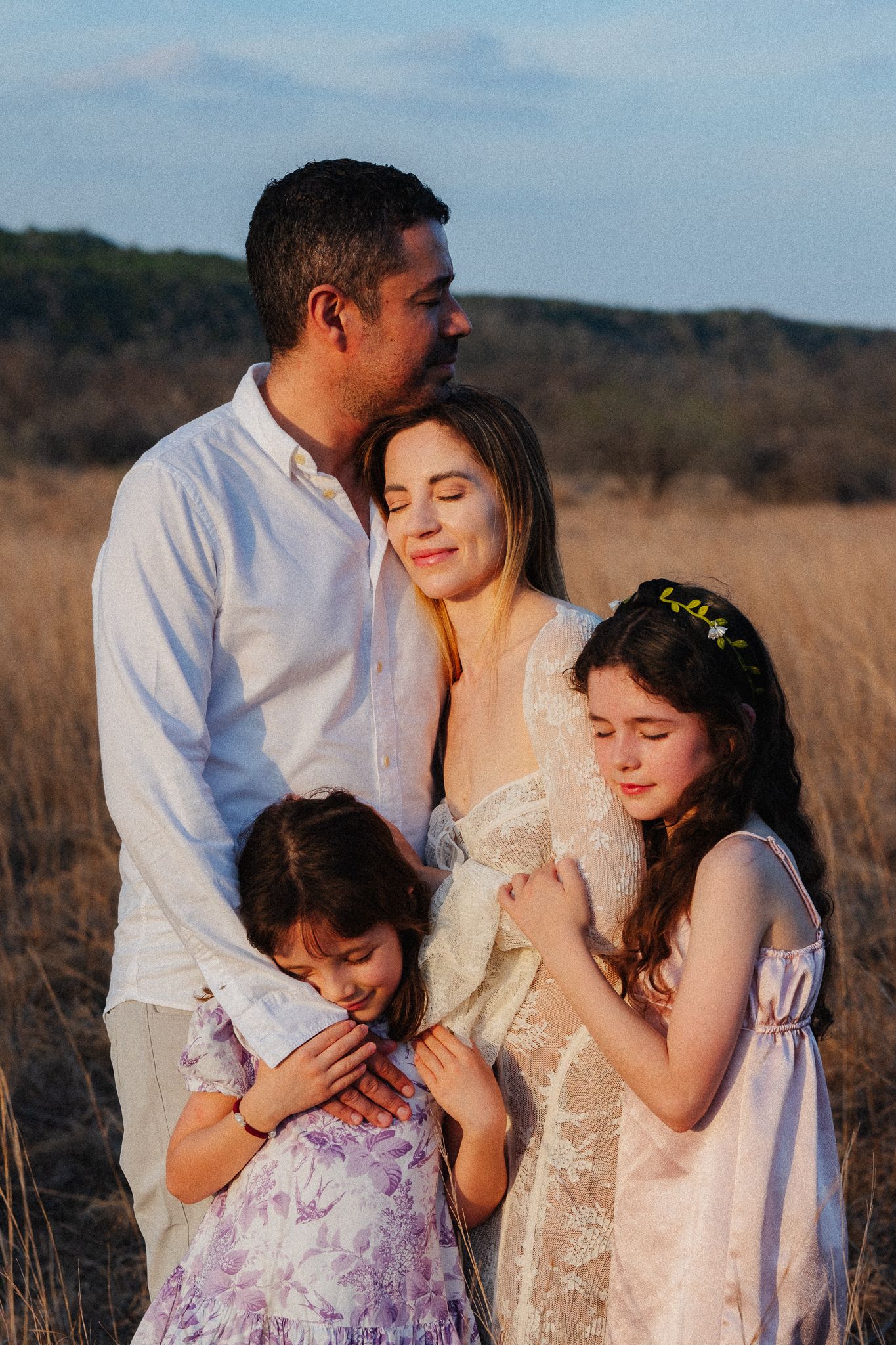 A family of four embraces in a golden field at sunset, with a pregnant mother, father, and two young daughters.