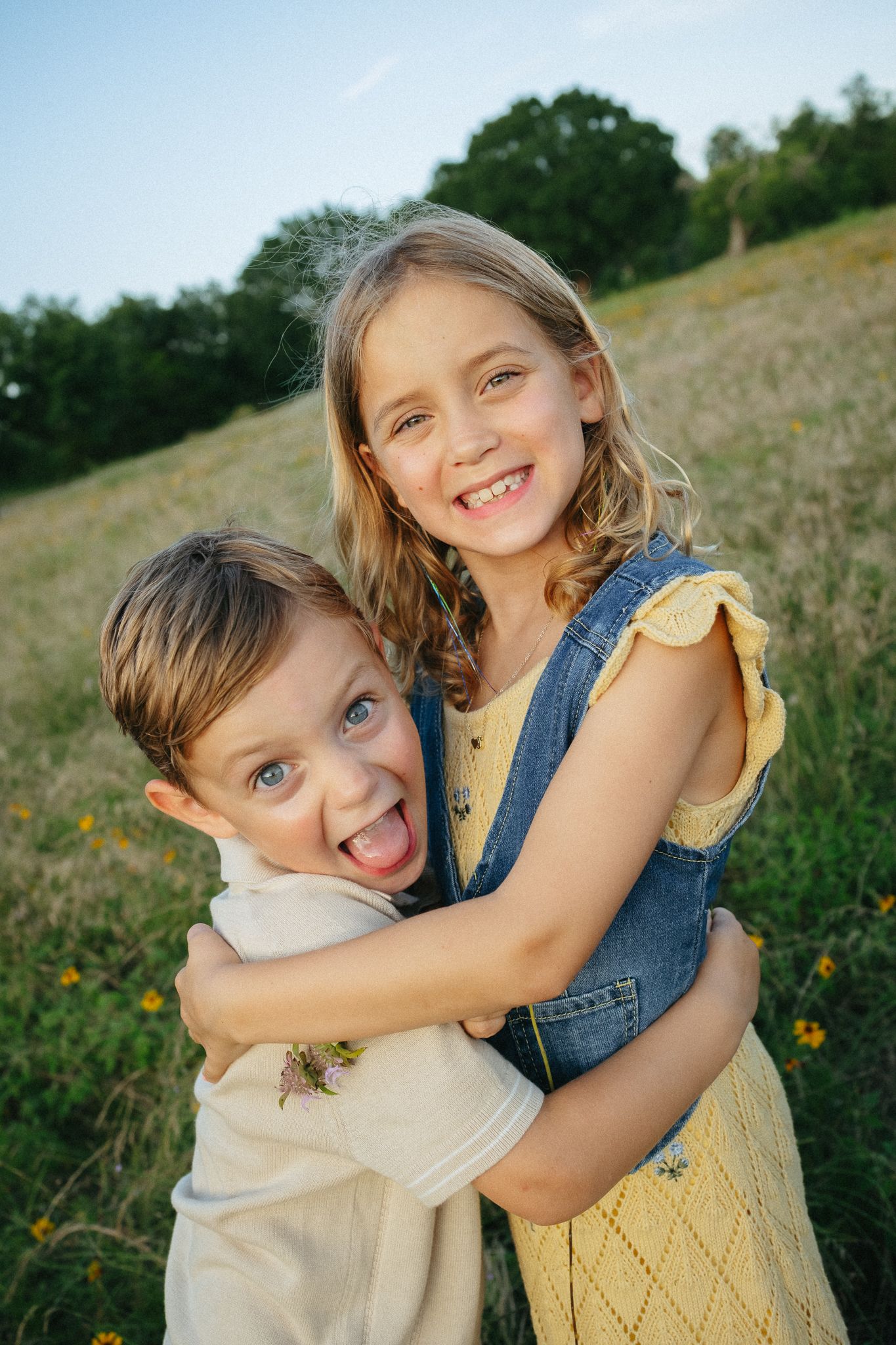 Two happy children hugging outdoors in a sunny meadow, girl in yellow dress smiling, boy sticking out tongue.