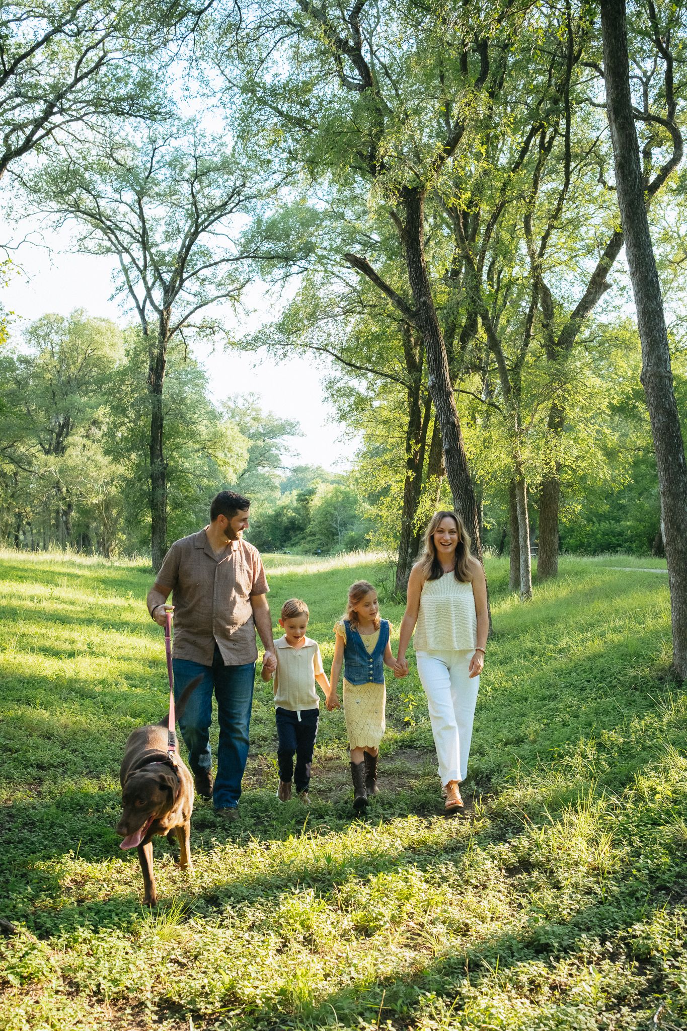 Family of four walking with a brown dog through a lush green park on a sunny day.