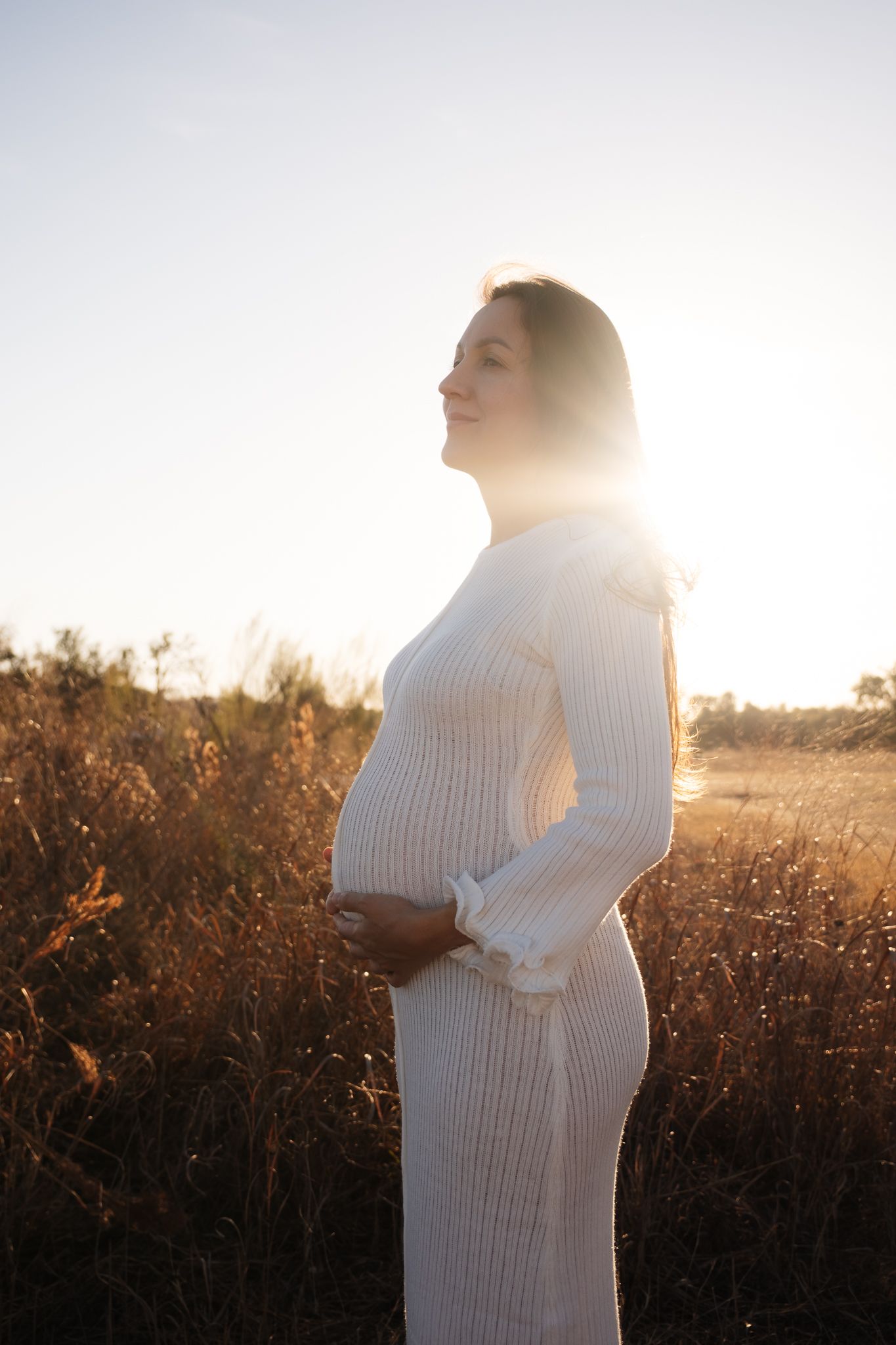 A pregnant woman in a white sweater dress stands in a golden field at sunset.