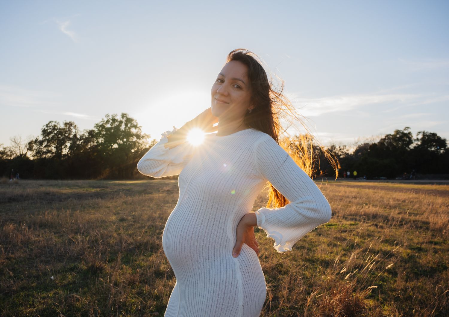 Silhouetted figure in white dress poses against sunset sky in rustic field setting.
