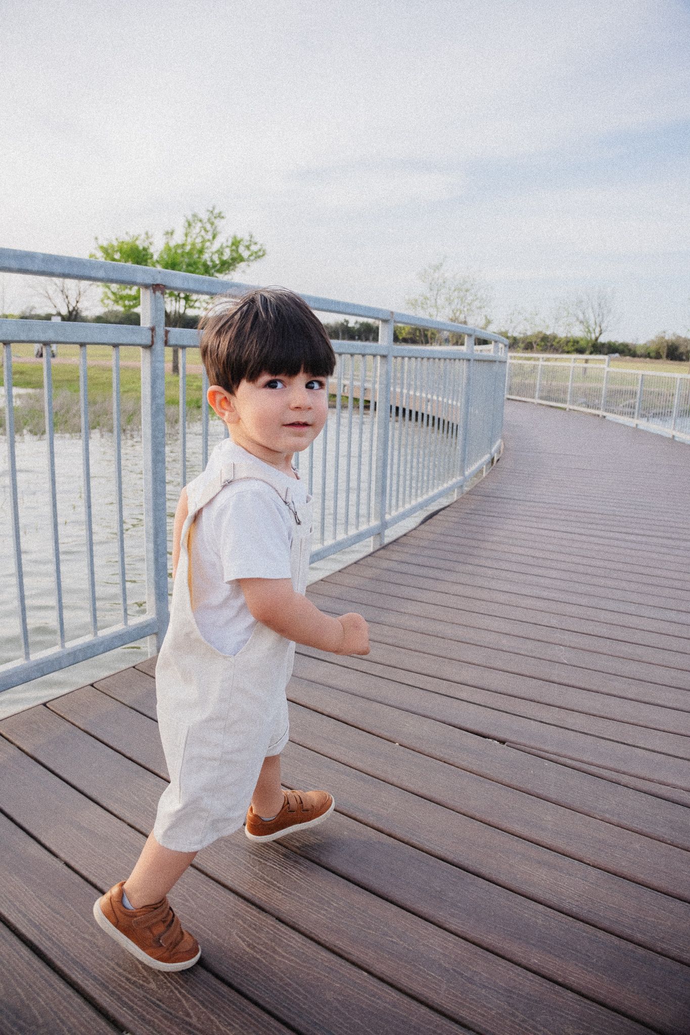 Small child wearing white overalls and brown shoes walks on wooden boardwalk with metal railing by the water.