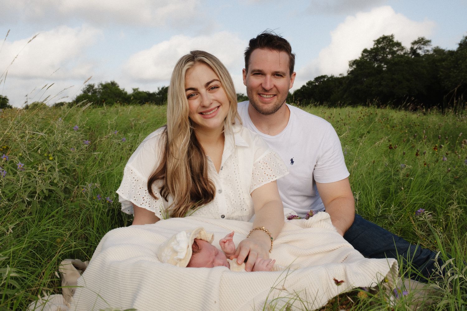 A couple sits together on a white blanket in a grassy field during a sunny outdoor photo session.