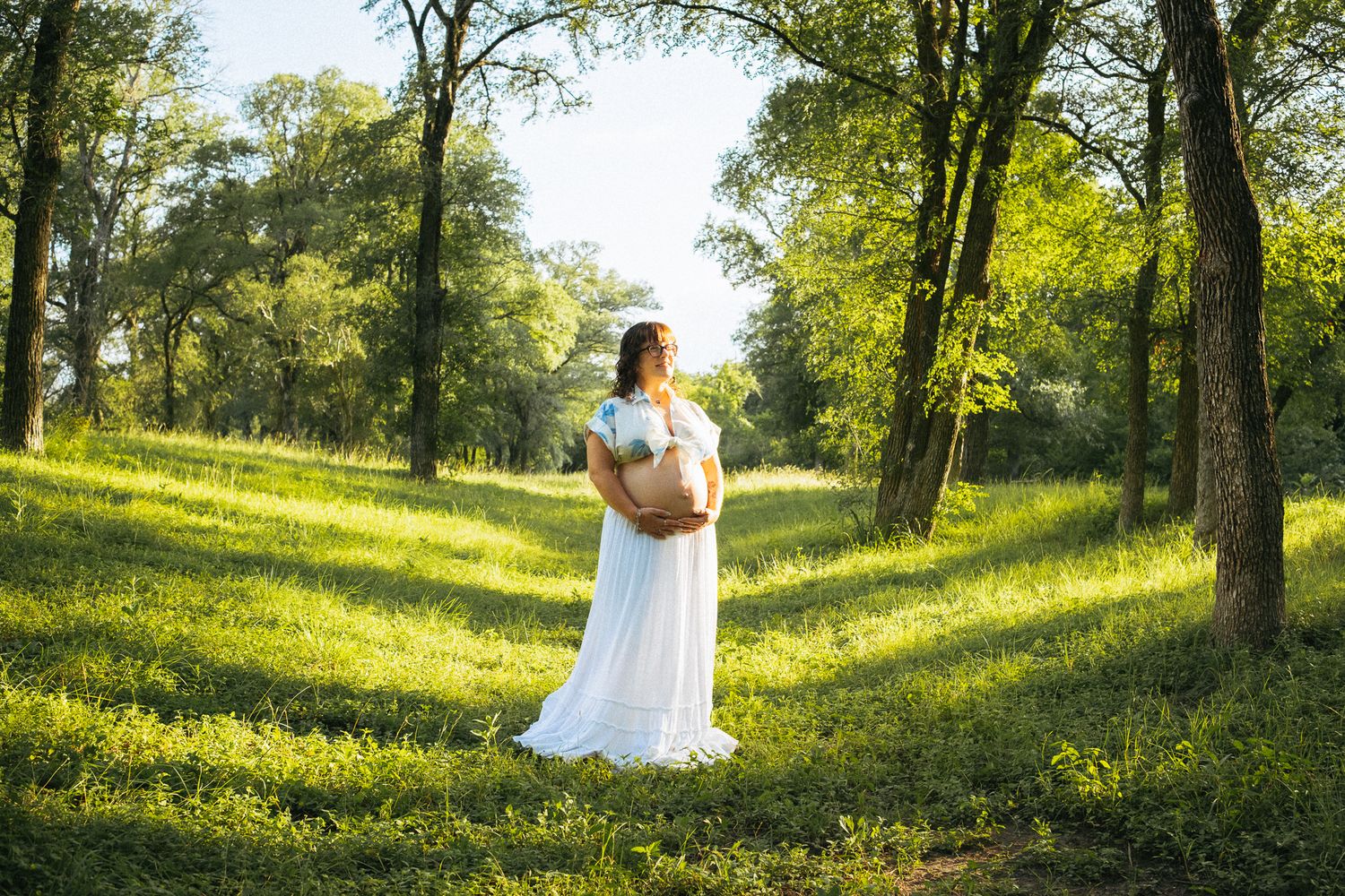 Woman in white dress holding a basket stands in a sunlit green forest clearing surrounded by tall trees.