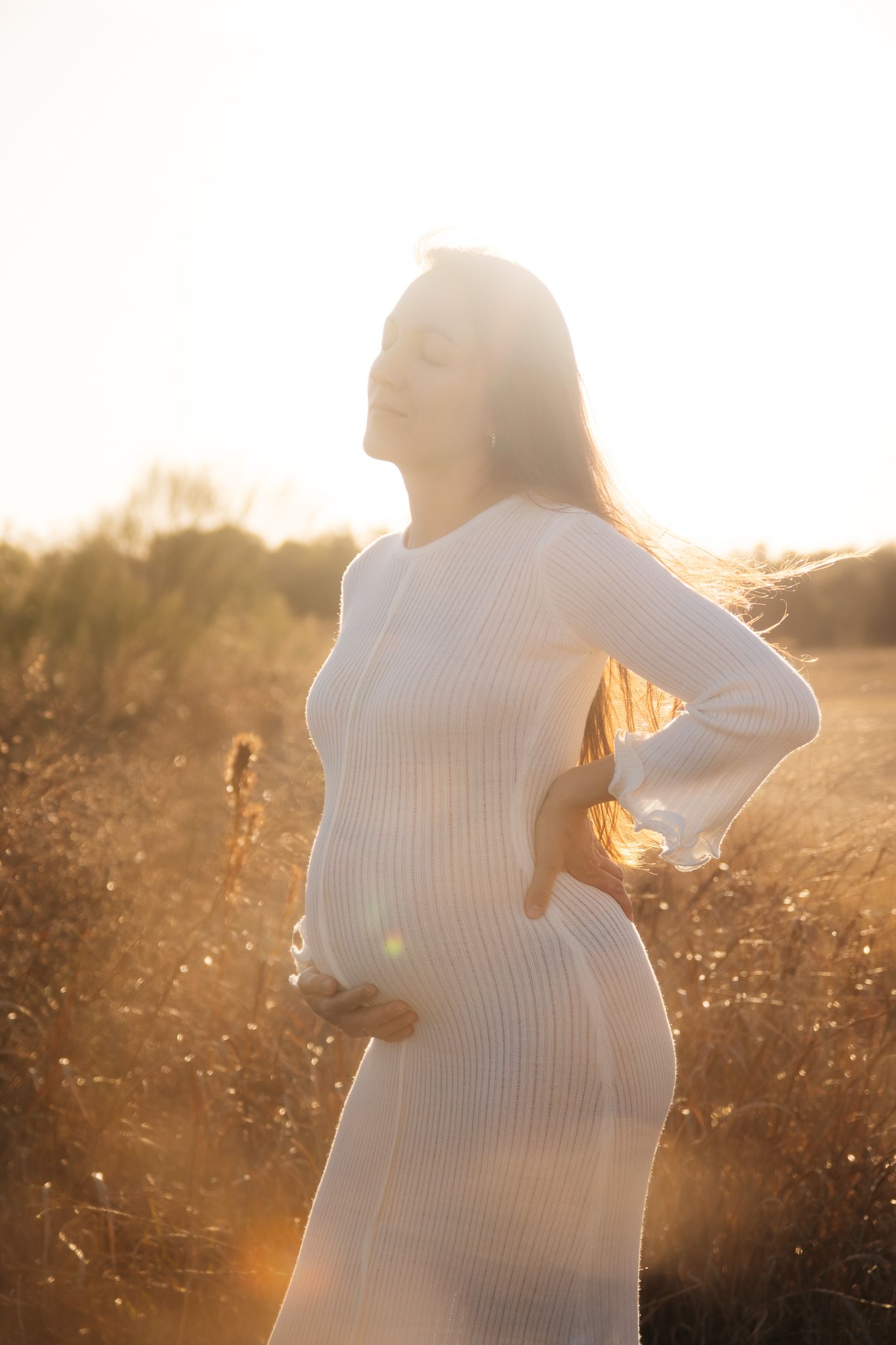 Silhouette of pregnant woman in white dress standing in golden sunlit field at sunset.