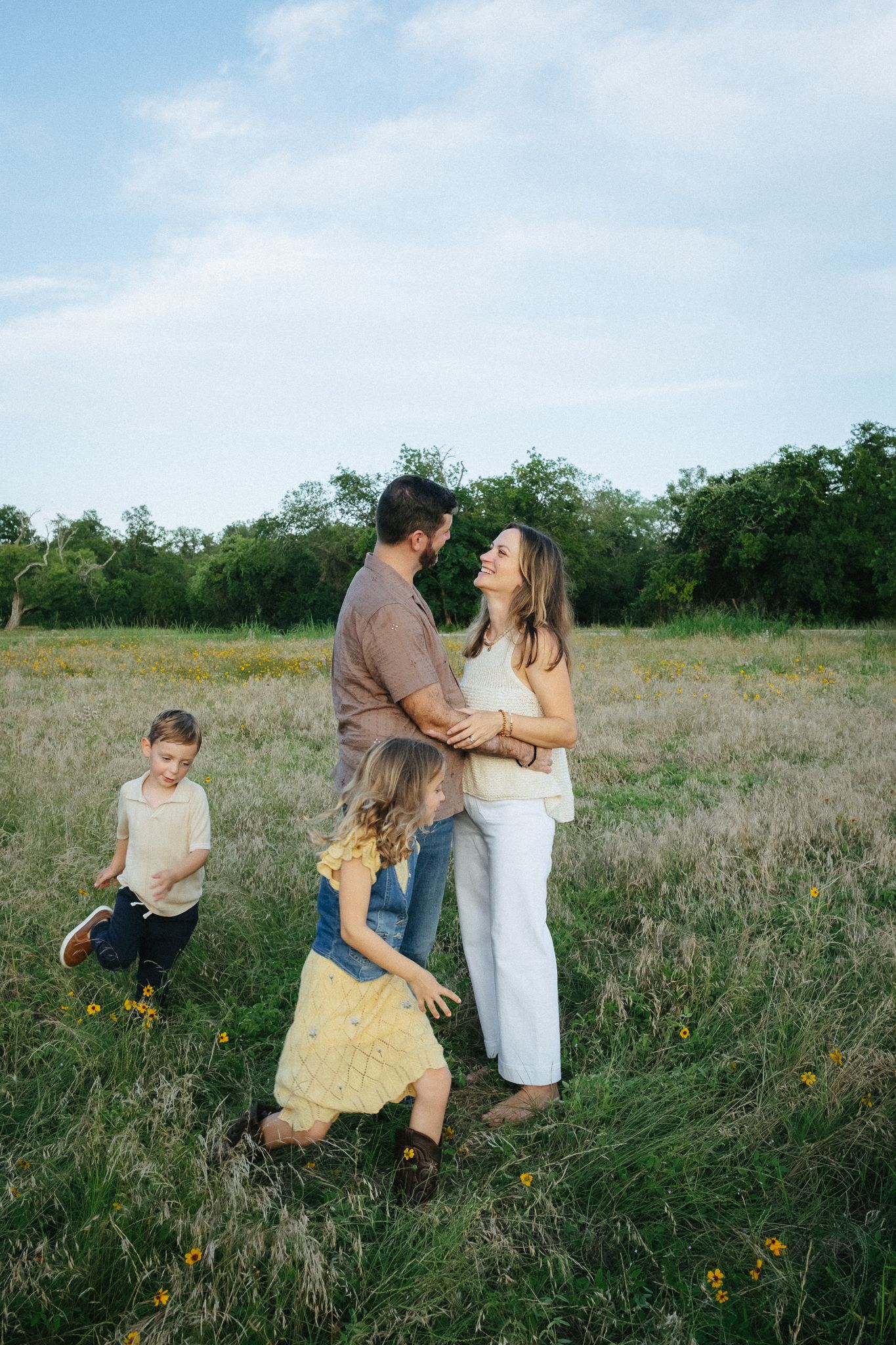 Family of four enjoying a sunny day in a wildflower meadow, parents embracing while children play around them.